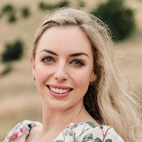 A smiling woman with blonde hair outdoors with a blurred natural background.