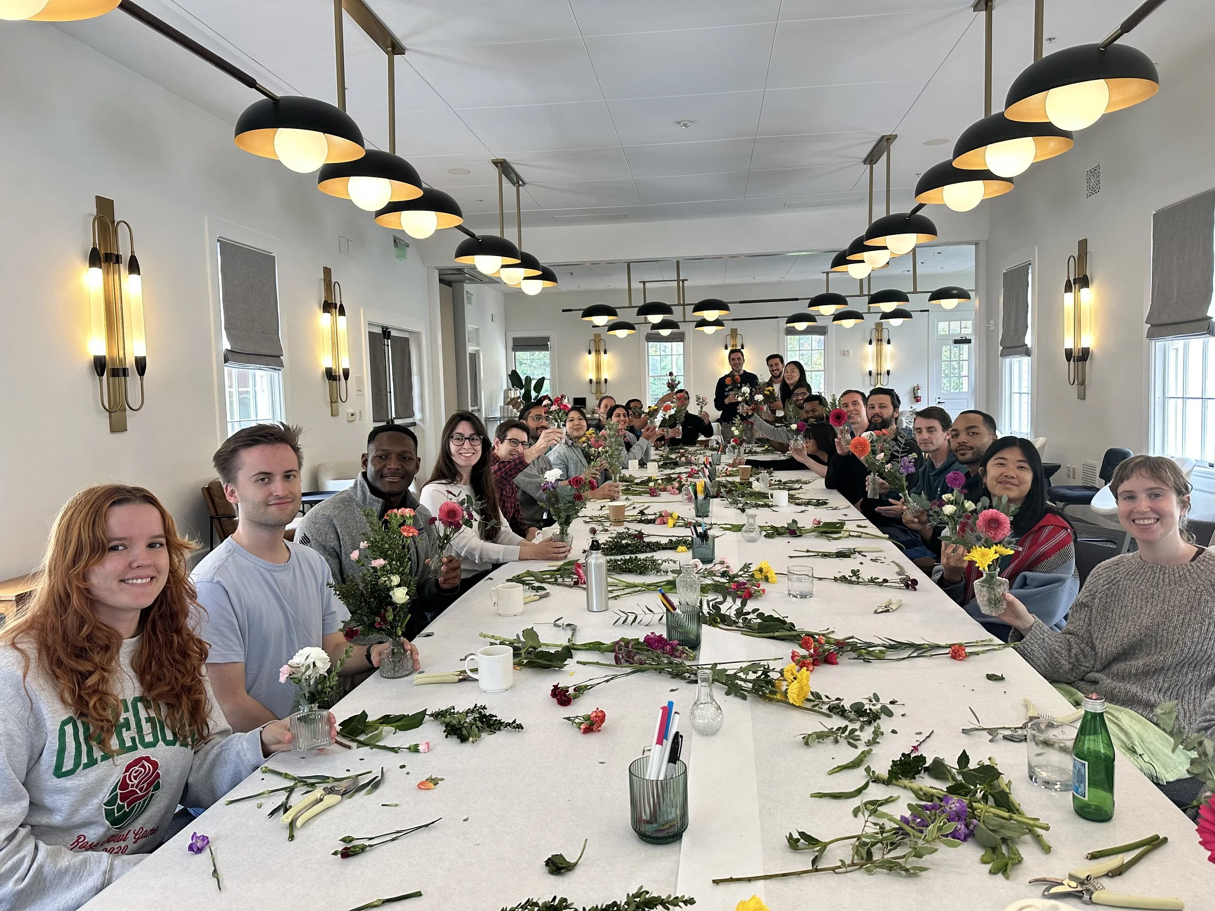 People sitting at a long table making floral arrangements in a bright room with modern lighting.