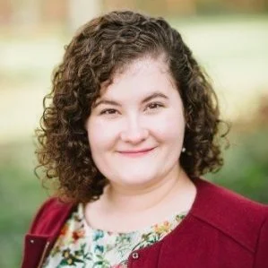 A woman with curly brown hair smiling outdoors, wearing a floral blouse and a maroon jacket.