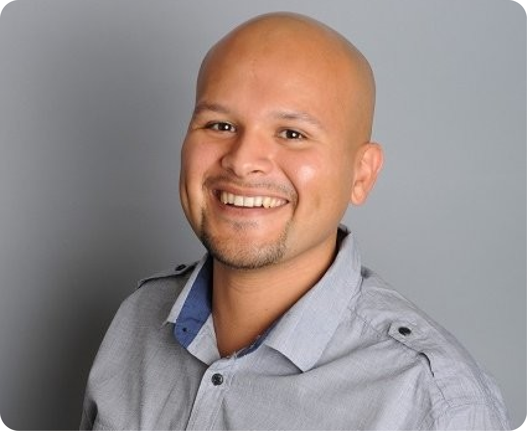 A smiling man with a shaved head, light skin, wearing a gray collared shirt with a blue inner lining, posing against a plain gray background.