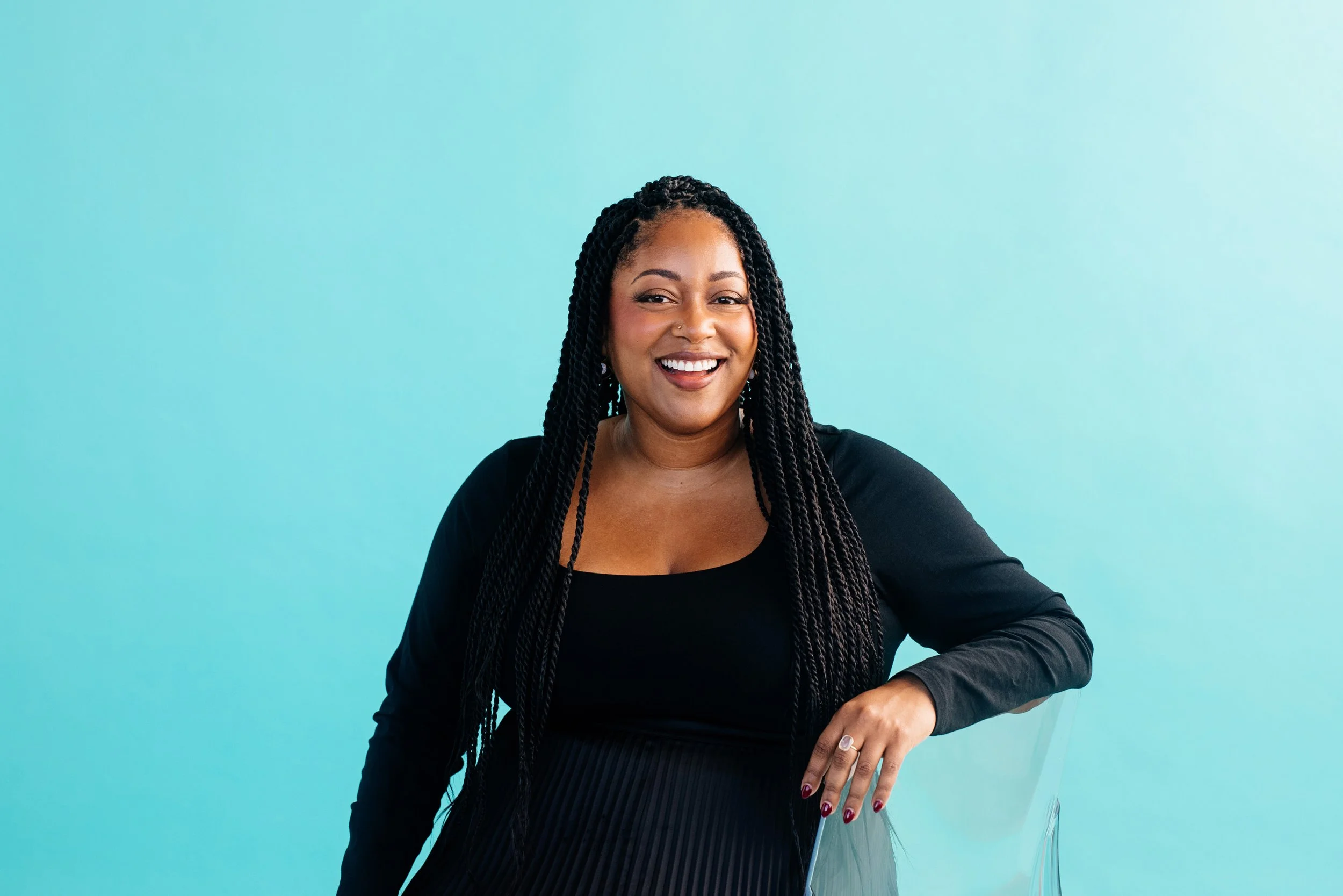A woman with long braided hair smiling, wearing a black top, against a light blue background.