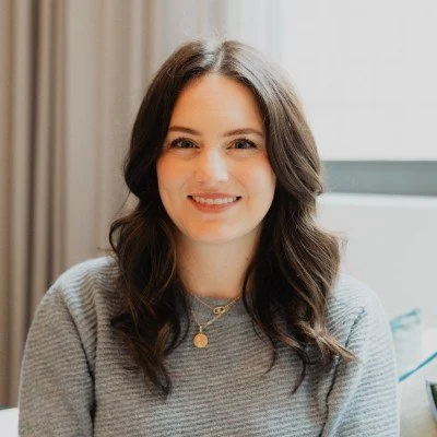 A young woman with long, wavy brown hair smiling at the camera, wearing a gray sweater and a delicate necklace, sitting indoors near a window with curtains.