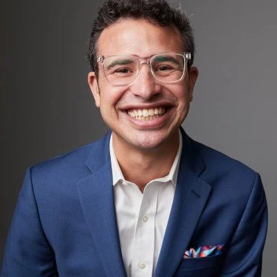 A smiling man with glasses wearing a blue suit and white shirt, standing against a gray background.