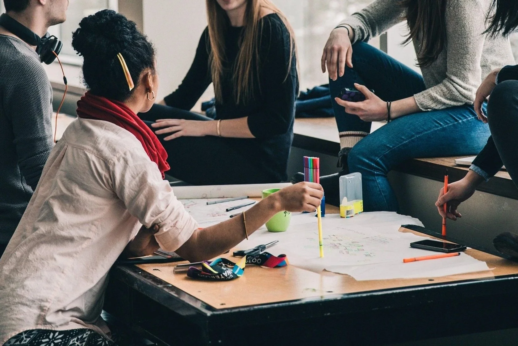 Group of young adults gathered around a table working on a creative project with markers, paper, and various art supplies in a casual indoor setting.