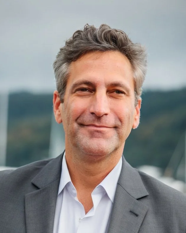 A middle-aged man with gray hair, wearing a gray suit and white shirt, smiling outdoors with a cloudy sky and blurred landscape or boats in the background.