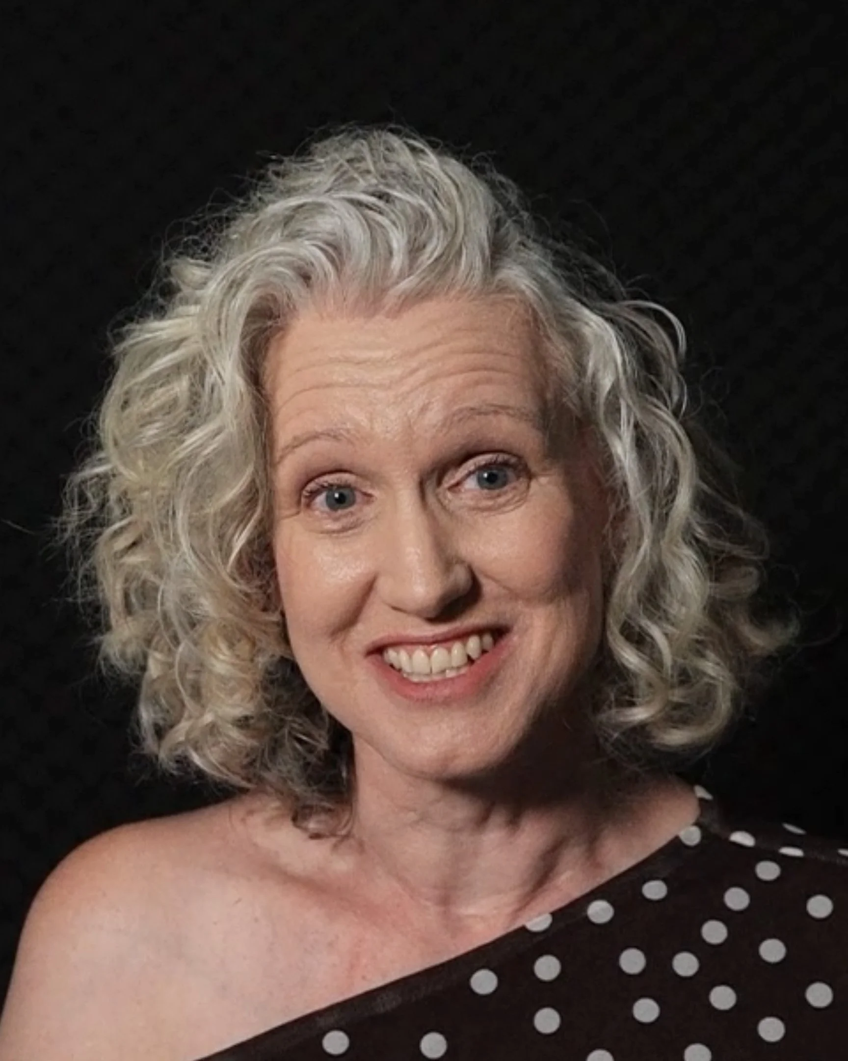 A woman with curly gray hair and blue eyes, smiling, wearing a black dress with white polka dots on one shoulder, against a black background.
