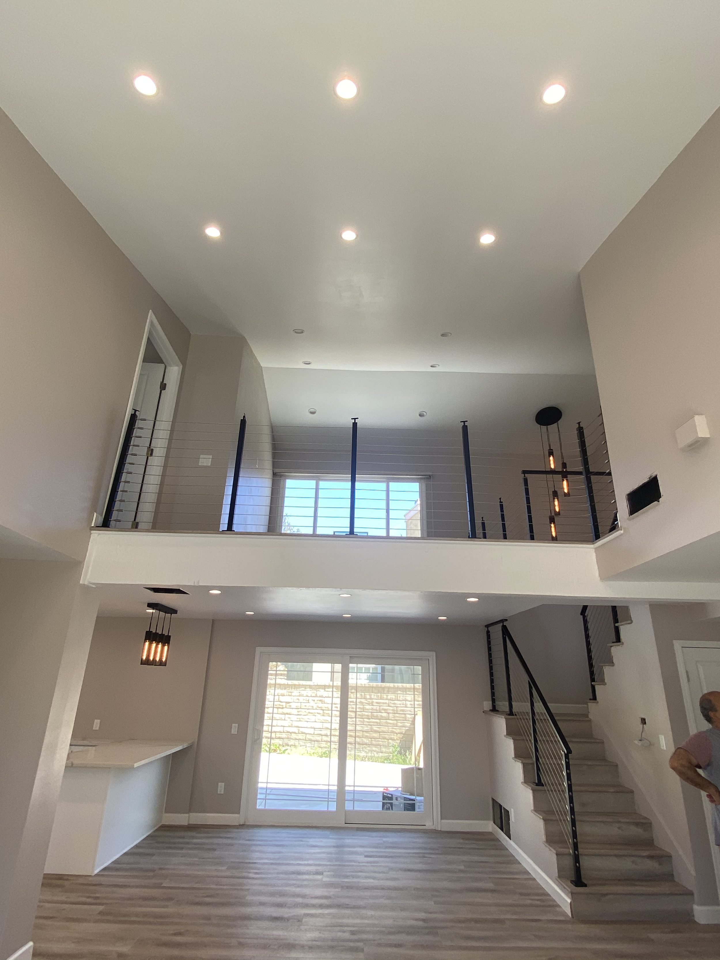 Interior view of a modern, spacious two-story house with an open floor plan. The lower level features a sliding glass door leading to an outdoor space, a staircase with black railing on the right, and a kitchen counter on the left. The second floor h