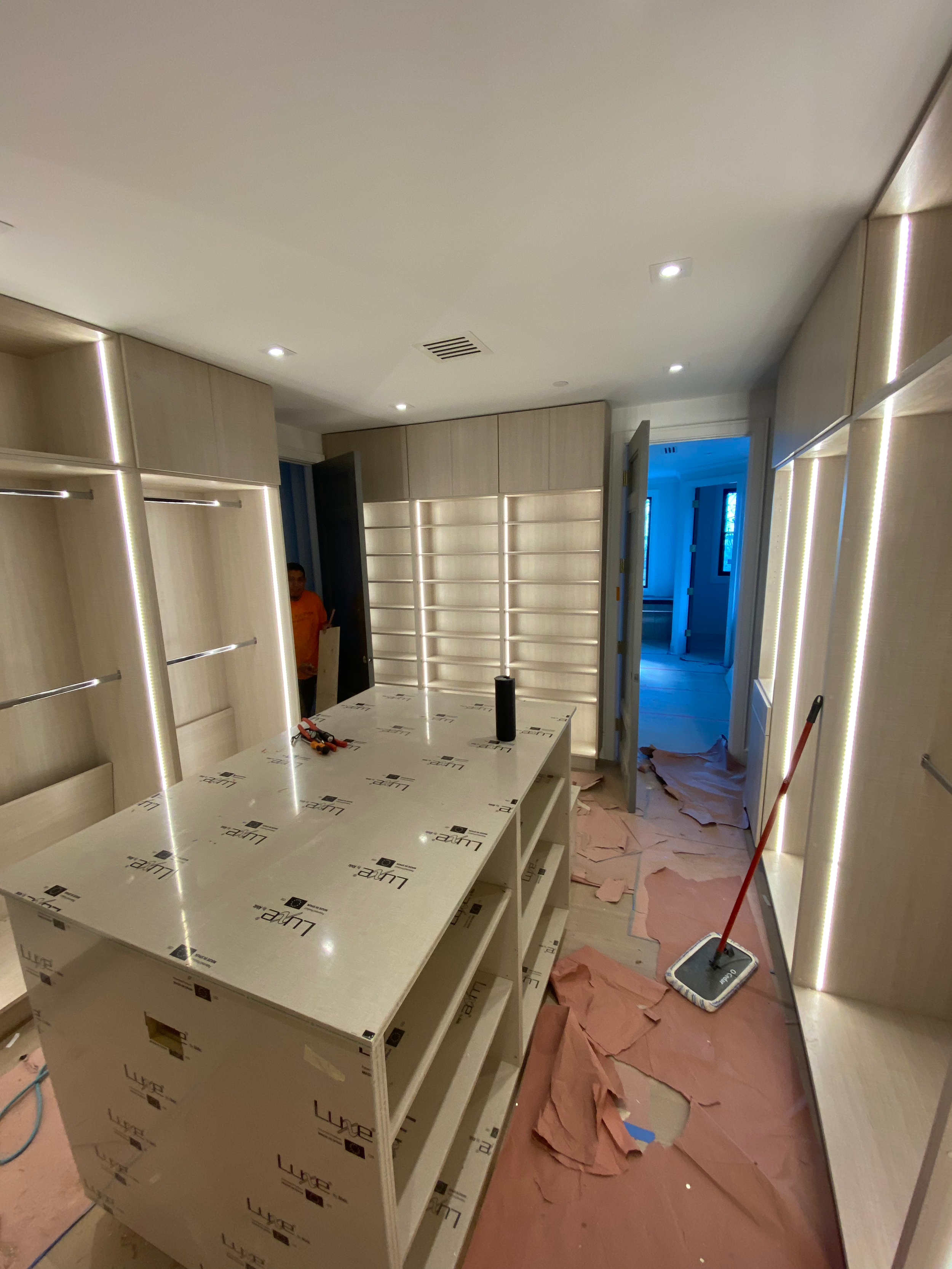 Interior of a room under construction with built-in wooden cabinets, a central workspace, construction tools, and cleaning equipment on the floor.