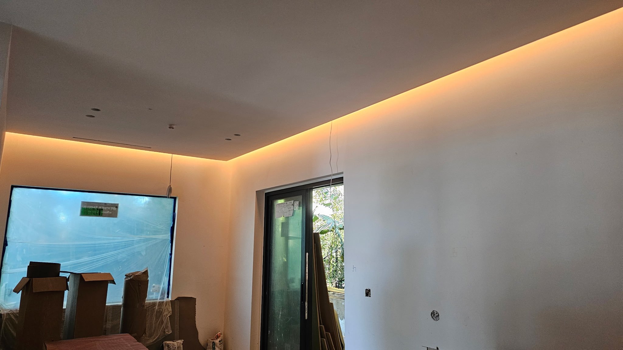 Interior of a room under construction with a white wall, a window covered with plastic, and a sliding door with a view of trees outside. There are boxes and construction materials on the floor and some wiring hanging from the ceiling.