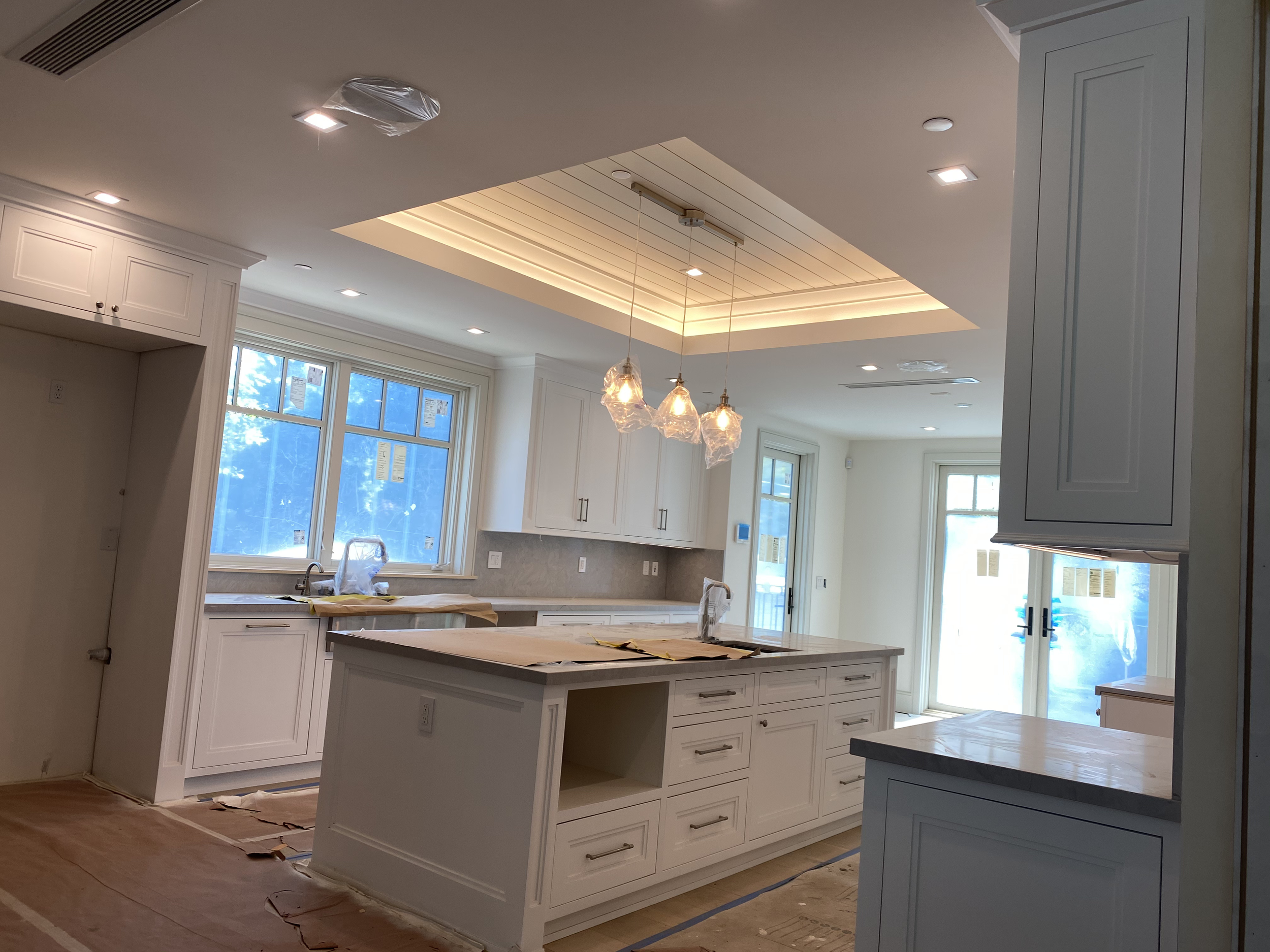 A modern white kitchen with an island, large windows, pendant lights, and recessed ceiling lights, in the process of being finished.