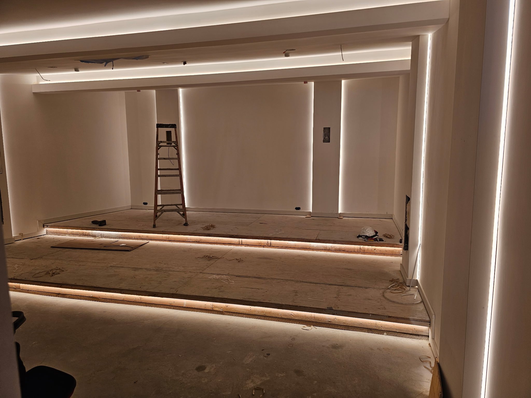 Under construction room with wooden flooring, drywall, LED strip lighting, ladder, and tools scattered around.