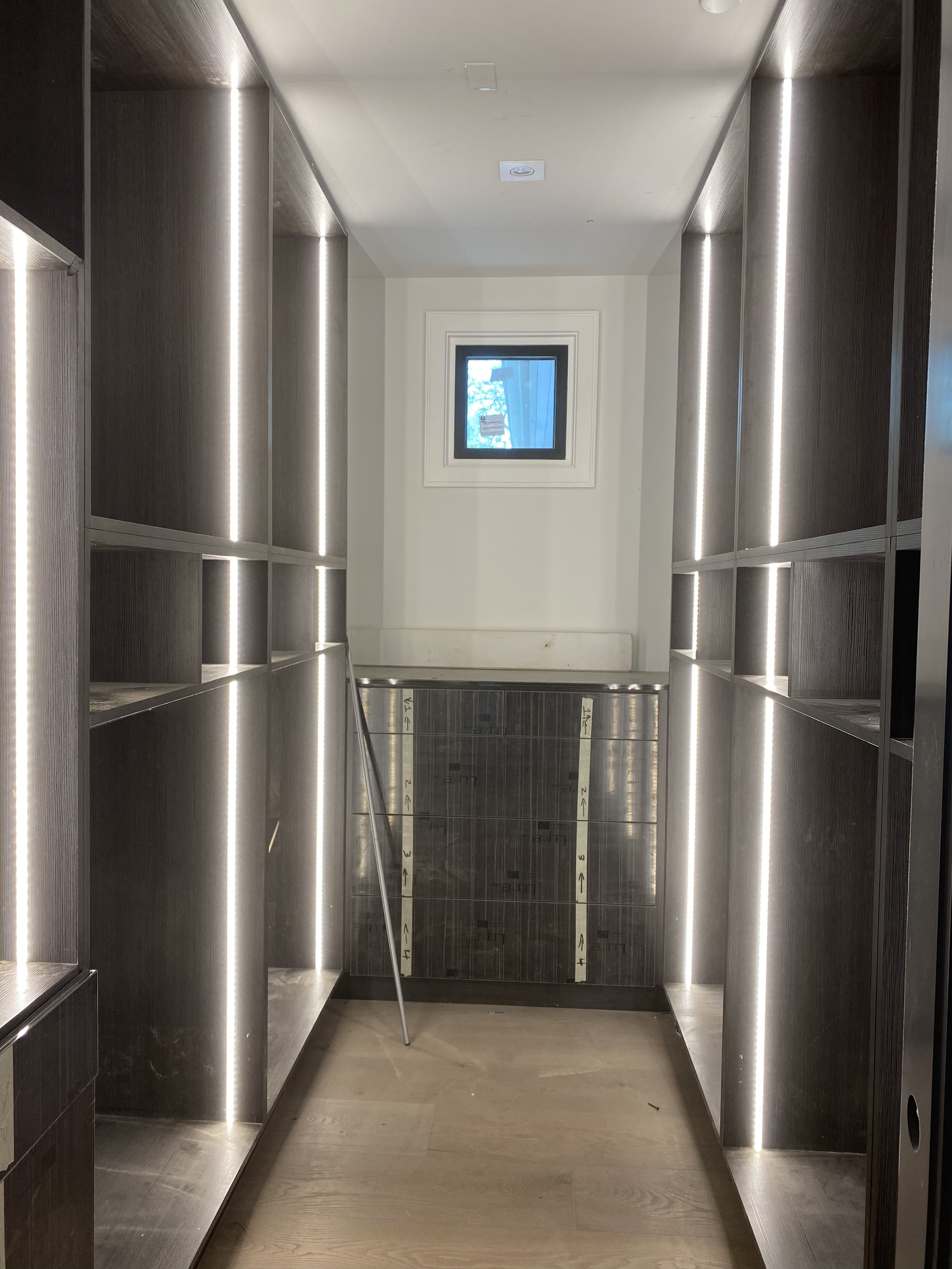 A small, narrow room with dark wooden shelving on both sides, LED strip lighting along the edges, a small window at the far end, a wooden floor, and a metal rod leaning against the back wall.