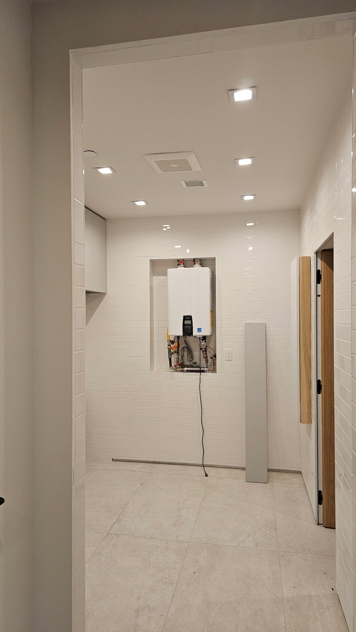 Unfinished utility room with white tiled walls, ceiling lights, and an installed tankless water heater with exposed pipes.