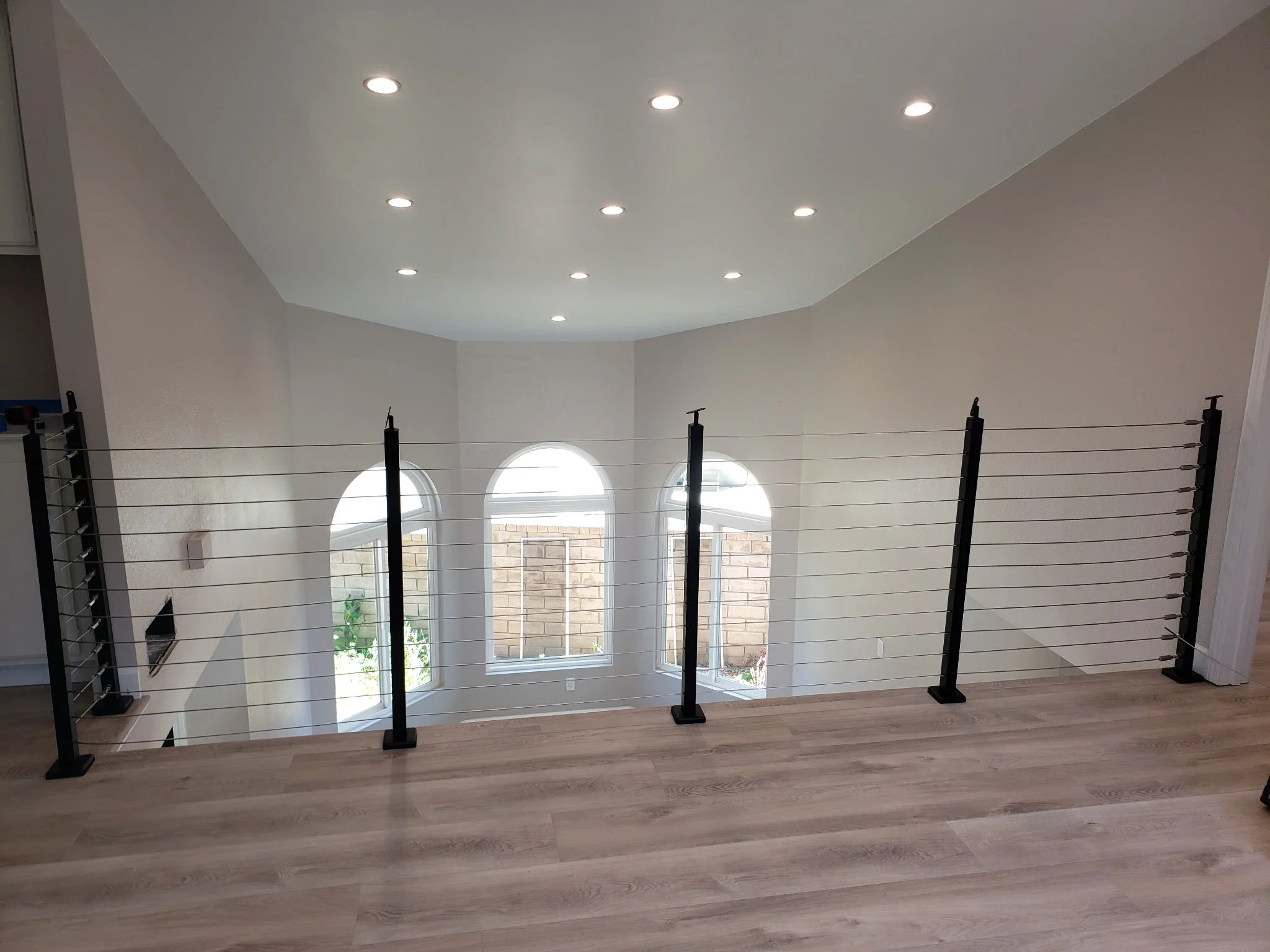 Interior view from the upper floor of a house, showing a safety barrier with black posts and horizontal cables overlooking a living room with three large arched windows and a white ceiling with recessed lights.