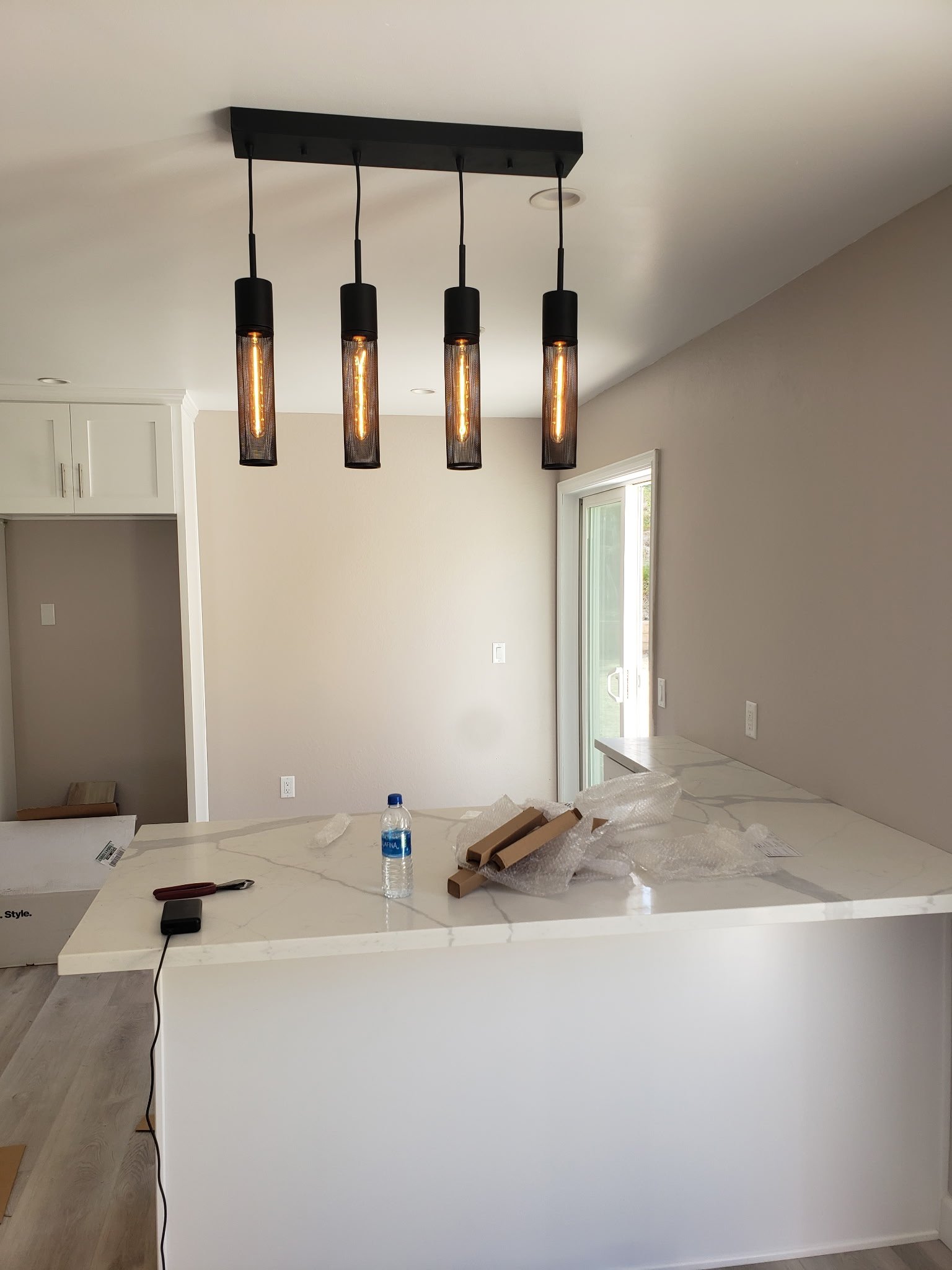 Modern kitchen with a white marble island, hanging black pendant lights, a bottle of water, and some packing materials on the counter. Sliding glass door leading outside.