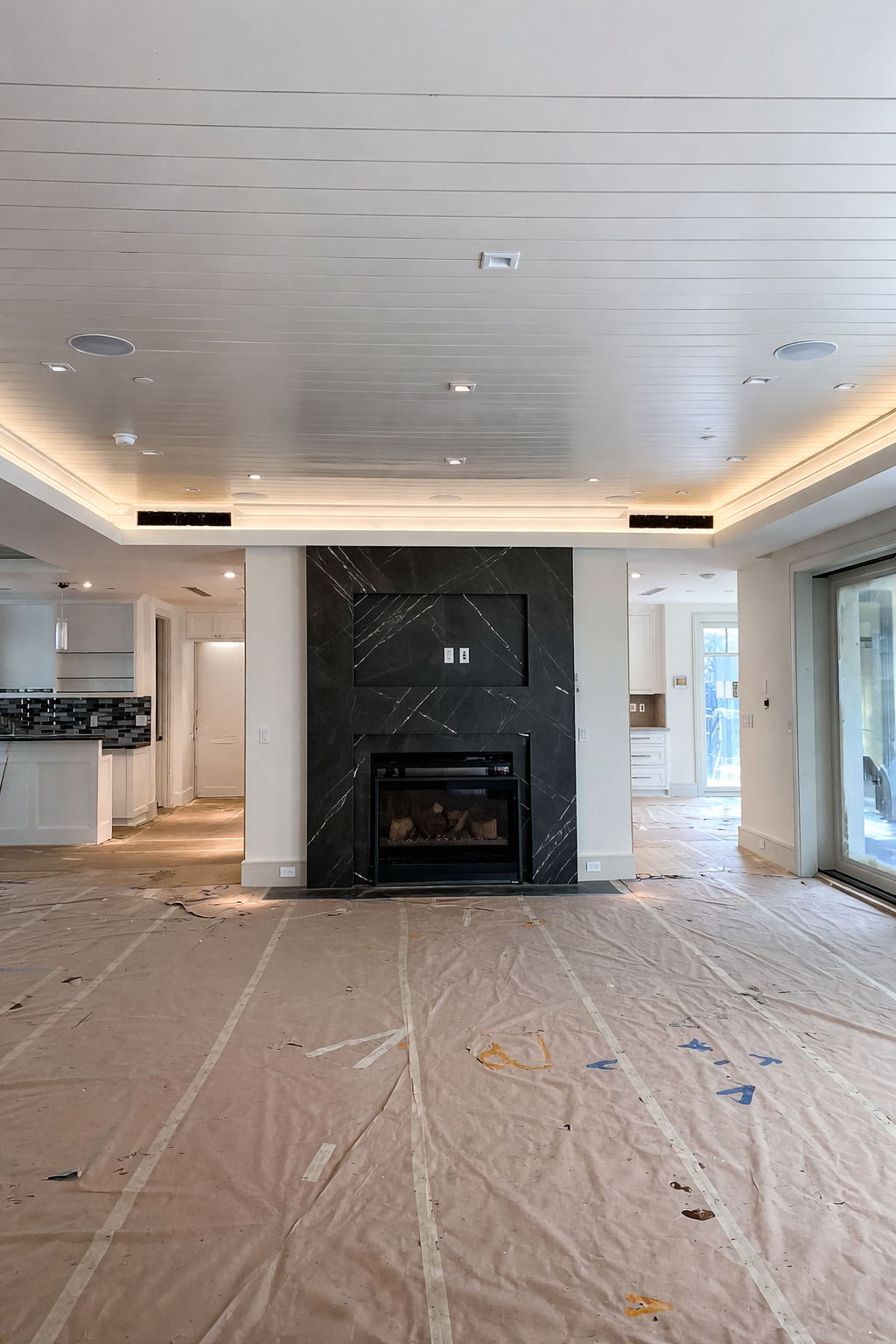Living room with a black marble fireplace, ceiling with recessed lighting, and large sliding glass doors, under construction with plastic covering on the floor.