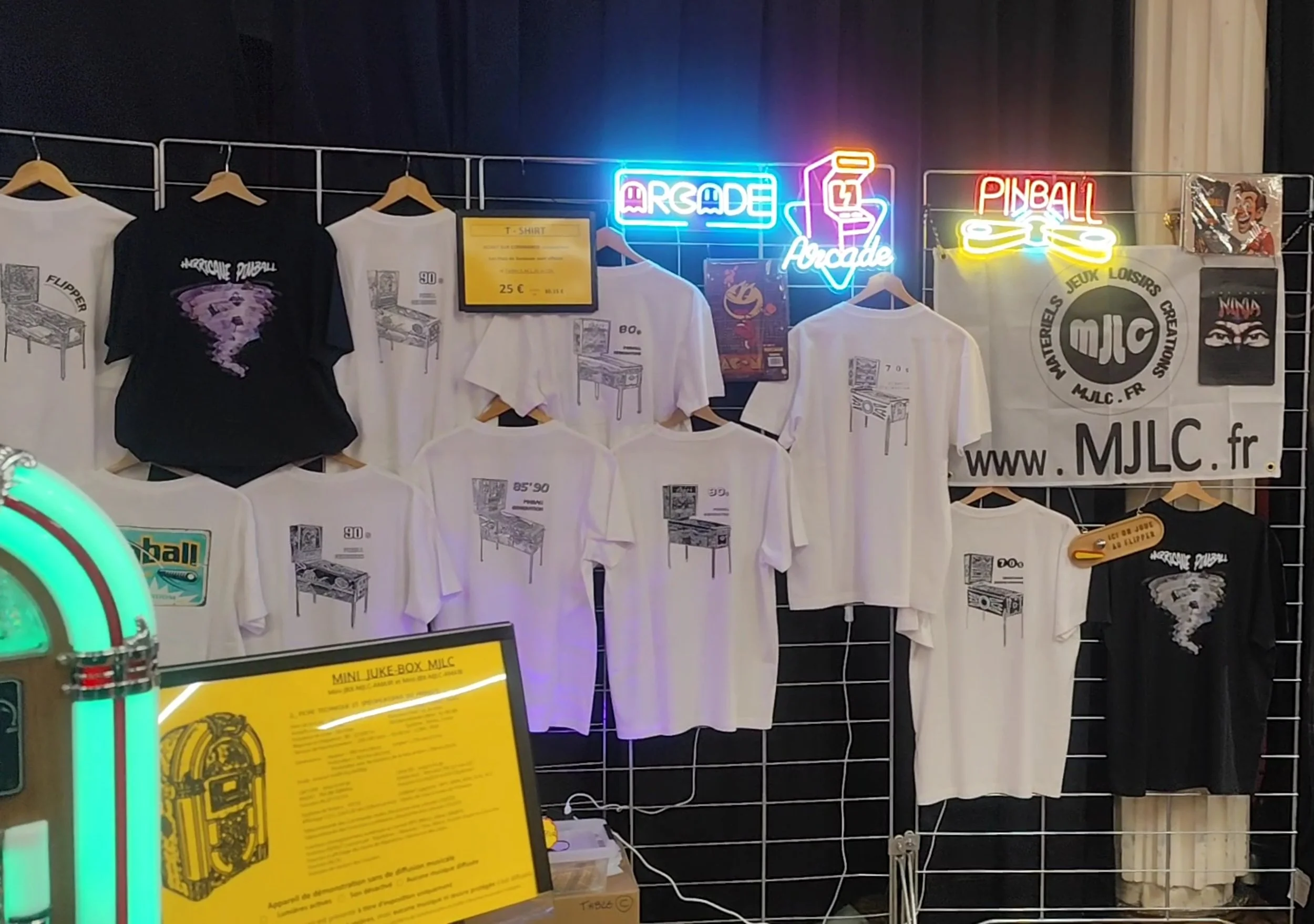 Vitrine avec des T-shirts avec des illustrations de machines à arcade rétro, néons en forme de : 'ARCADE', 'Pinball' et un logo MJLC, fond noir et grille métallique, panneau d'affichage jaune avec prix 25 €, bandeau publicitaire MJLC et décoration av