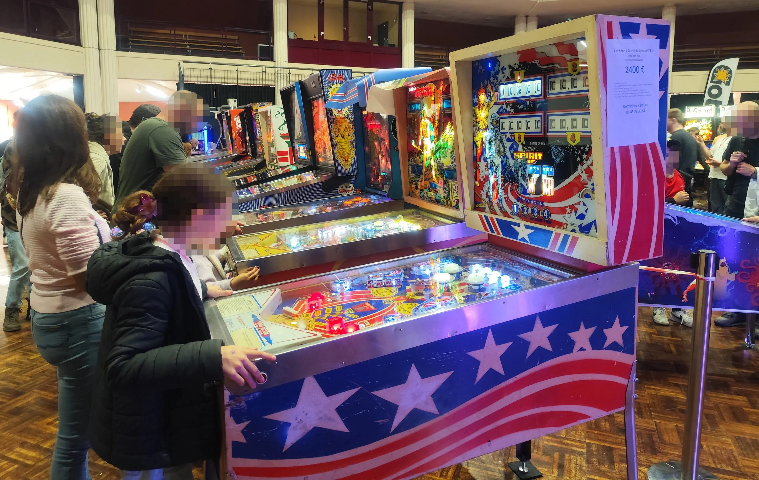 Des personnes jouent aux flippers dans une salle de jeux (le salon du flipper d'Ambérieu-en-Bugey). L'installation de flipper est décorée avec un thème patriotique américain, contenant des étoiles et des couleurs rouge, blanc et bleu.