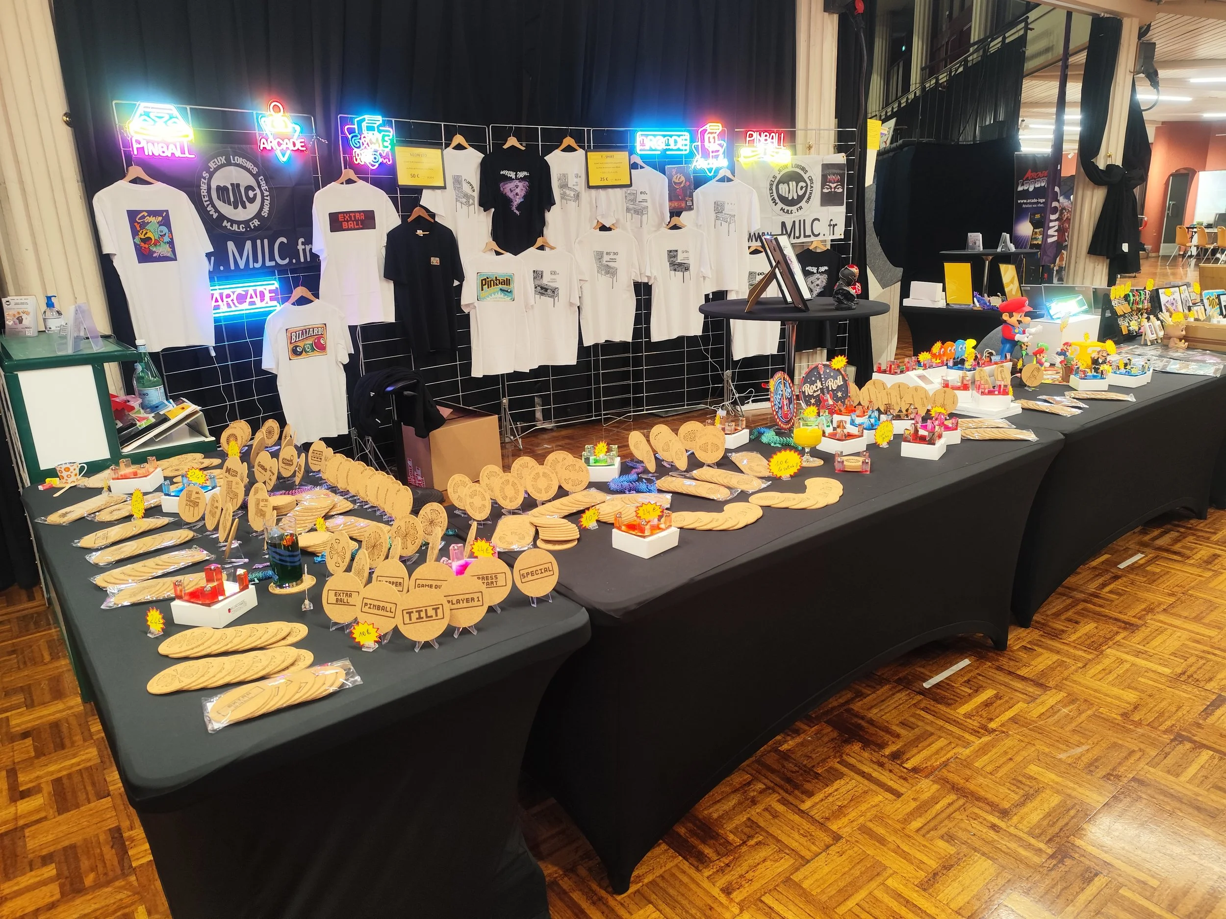 Un stand de vente d'articles liés au jeu vidéo arcade, pinball et flipper, avec des t-shirts, des figurines de Mario Bros, des décorations, et des plaques en bois et métal avec des inscriptions sur le thème du jeu automatique de café, stand mjlc.fr. 