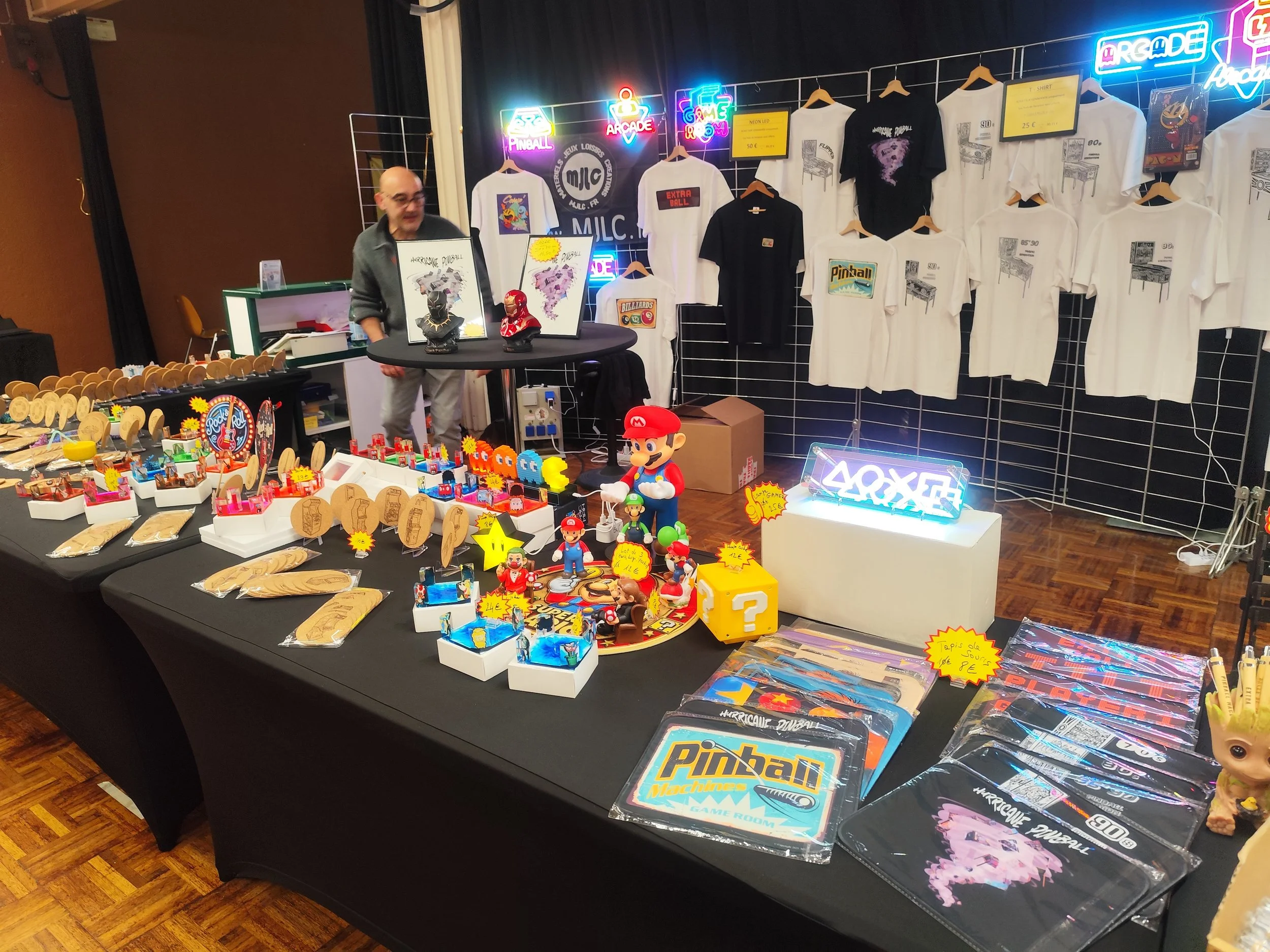 Vente d'objets liés aux jeux rétro et vintage vidéo arcade et flipper, avec t-shirts, figurines, décorations originales et accessoires dans le salon du flipper d'Ambérieu en Bugey en 2025.