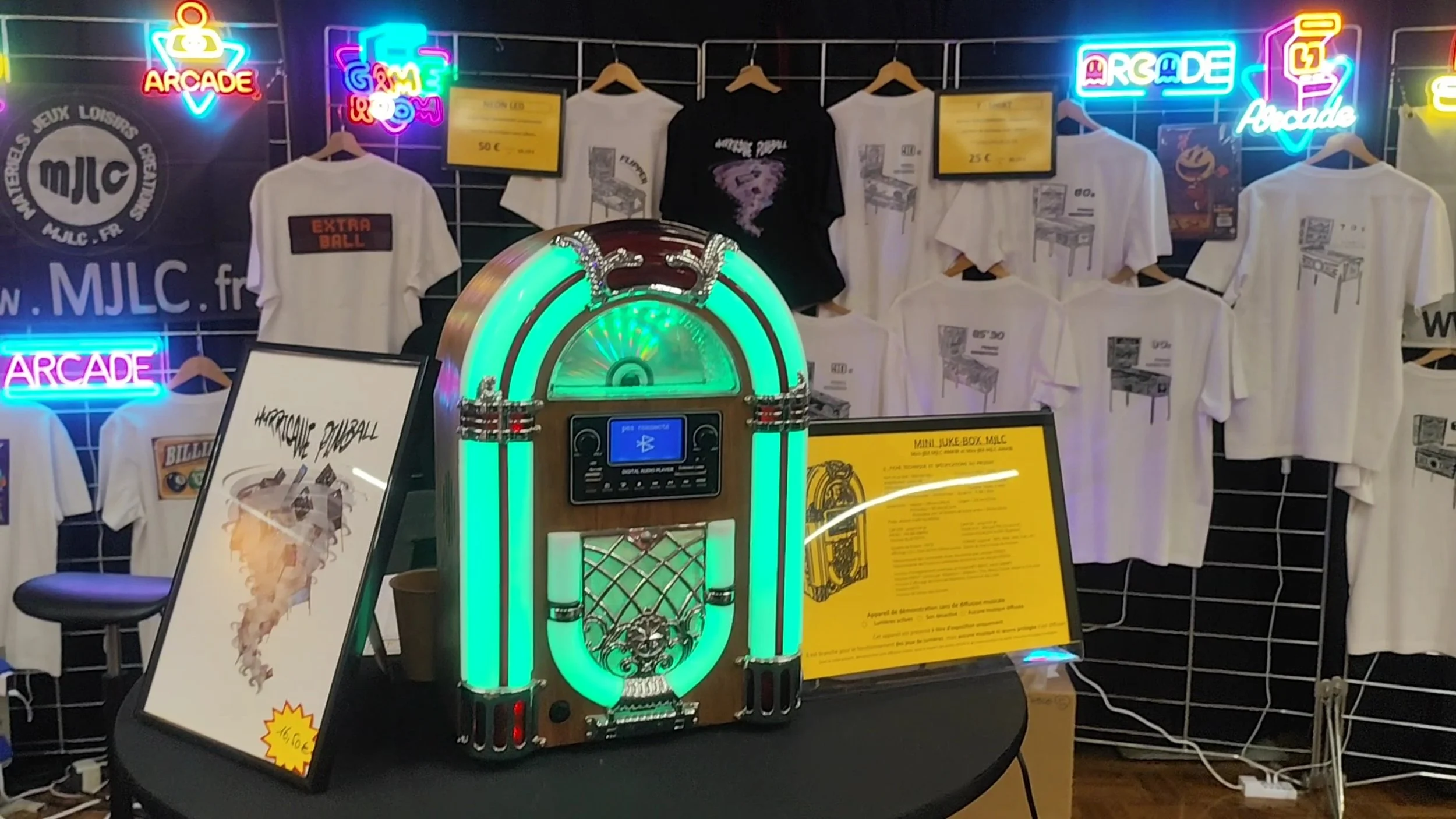 Accueil d'une boutique avec une guitare-jukebox rétro en premier plan, derrière un mur noir avec des t-shirts imprimés et des néons bleu, rose, vert, jaune affichant 'ARCADE' et 'GAME & ROOM'.