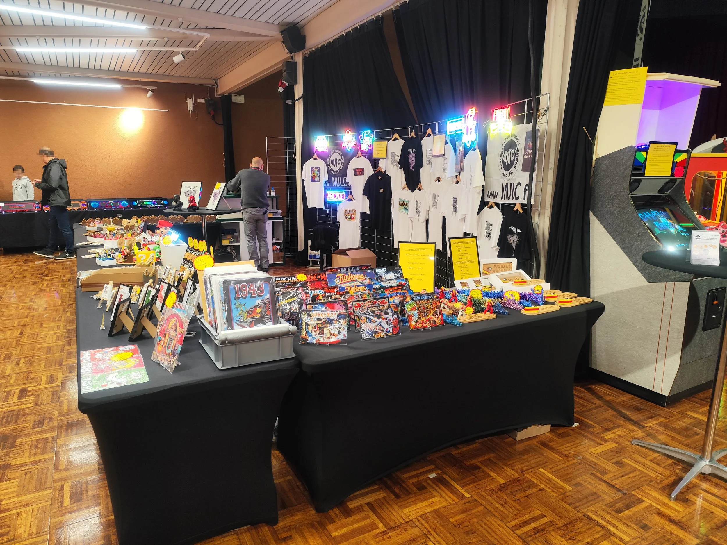 photo du stand de mjlc.fr spécialisé dans le jeu de café, affichant des articles de collection et déco, des plaques murales, des figurines, des t-shirts et d'autres objets de décoration rétro et moderne, ainsi que des jeux d'arcade en arrière-plan.