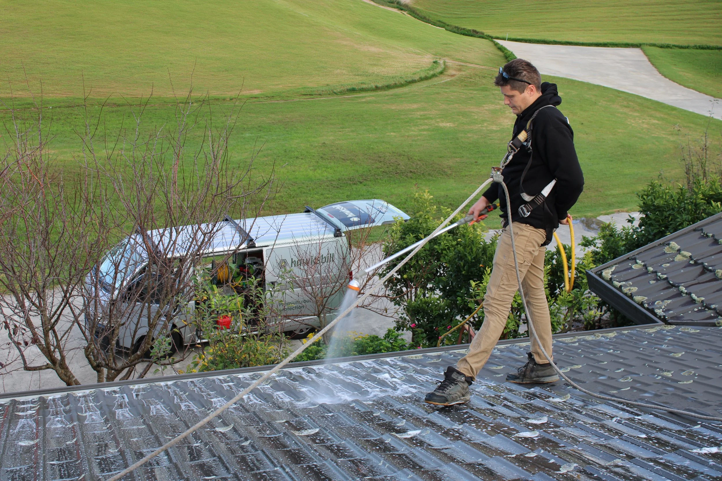A man is cleaning a metal rooftop with a hose, wearing a harness and standing on the roof's edge. There is a van parked on a driveway below, with a grassy landscape extending into the distance.