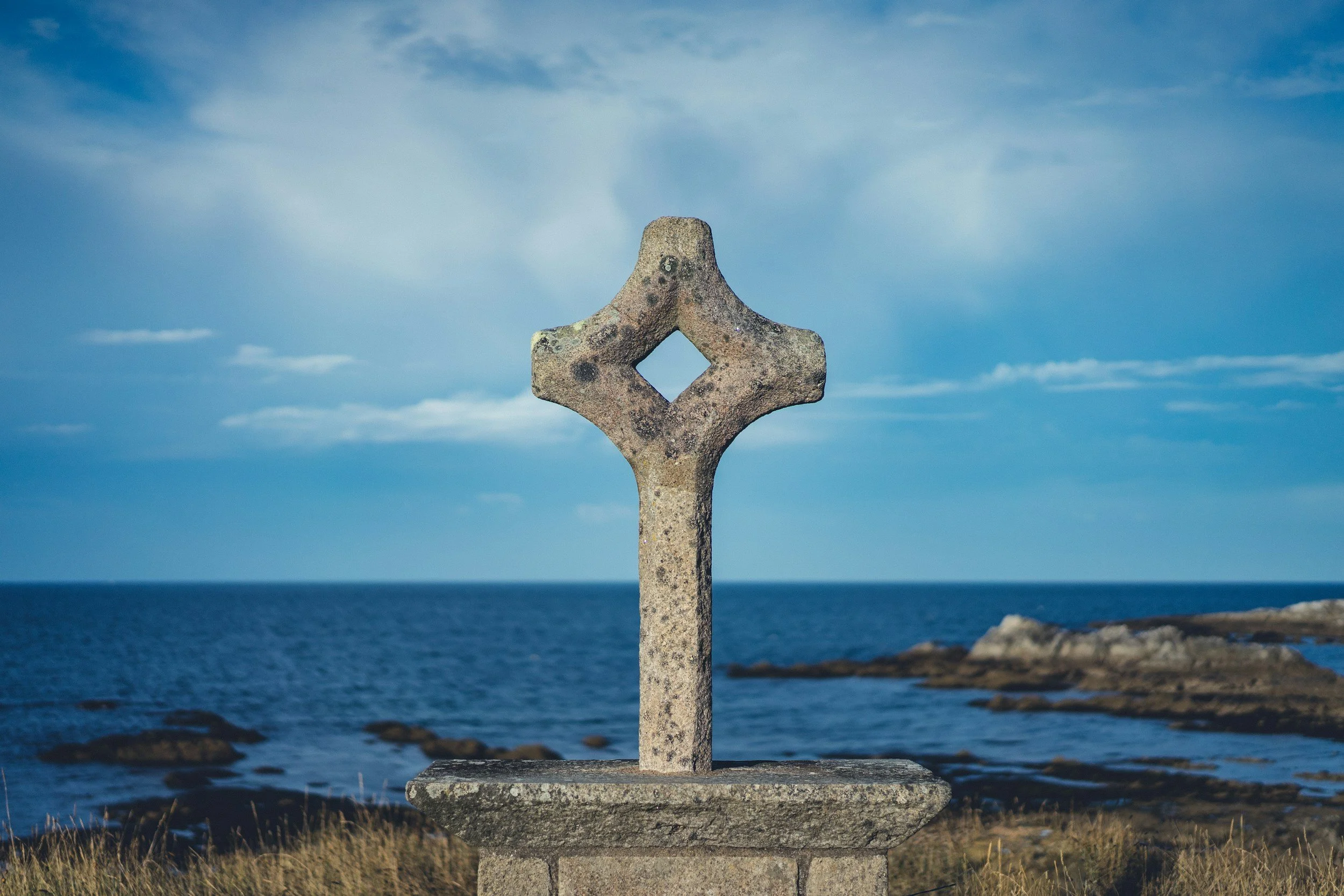 A stone cross monument on a coastal landscape with rocks and the ocean in the background under a partly cloudy sky.