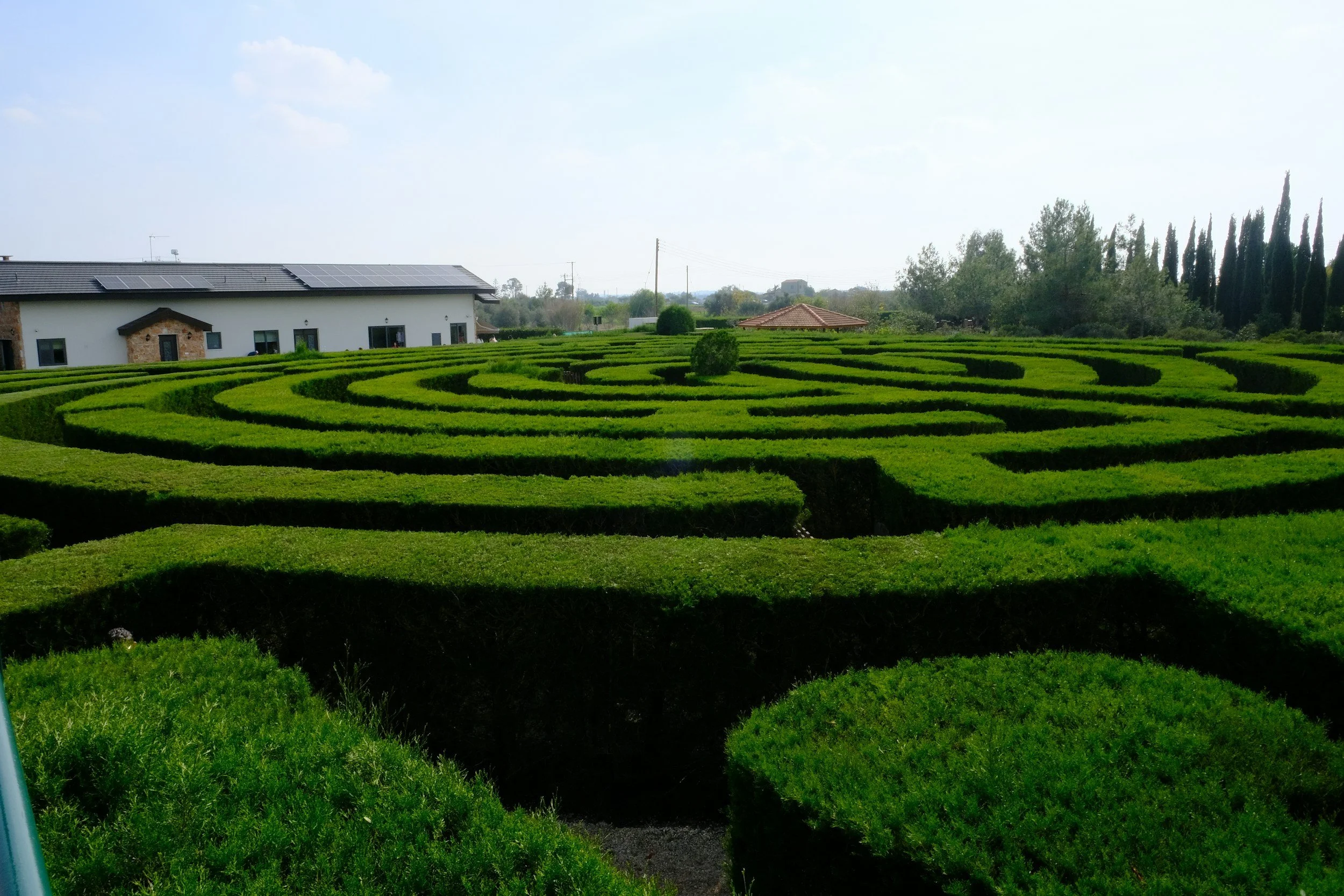 A maze made of neatly trimmed green hedges in front of a white building with solar panels on the roof, surrounded by trees and a cloudy sky.