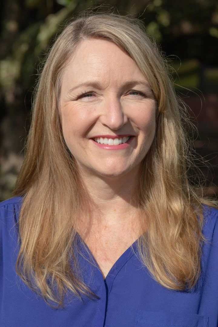 A smiling woman with long blonde hair wearing a blue shirt outdoors in natural sunlight.