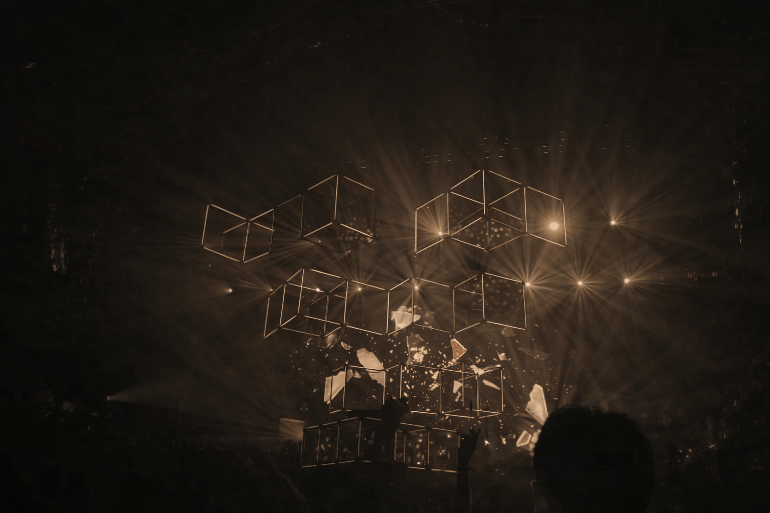 A dark stage with geometric cube structures emitting light, surrounded by sparks and beams, creating a visually striking light show.