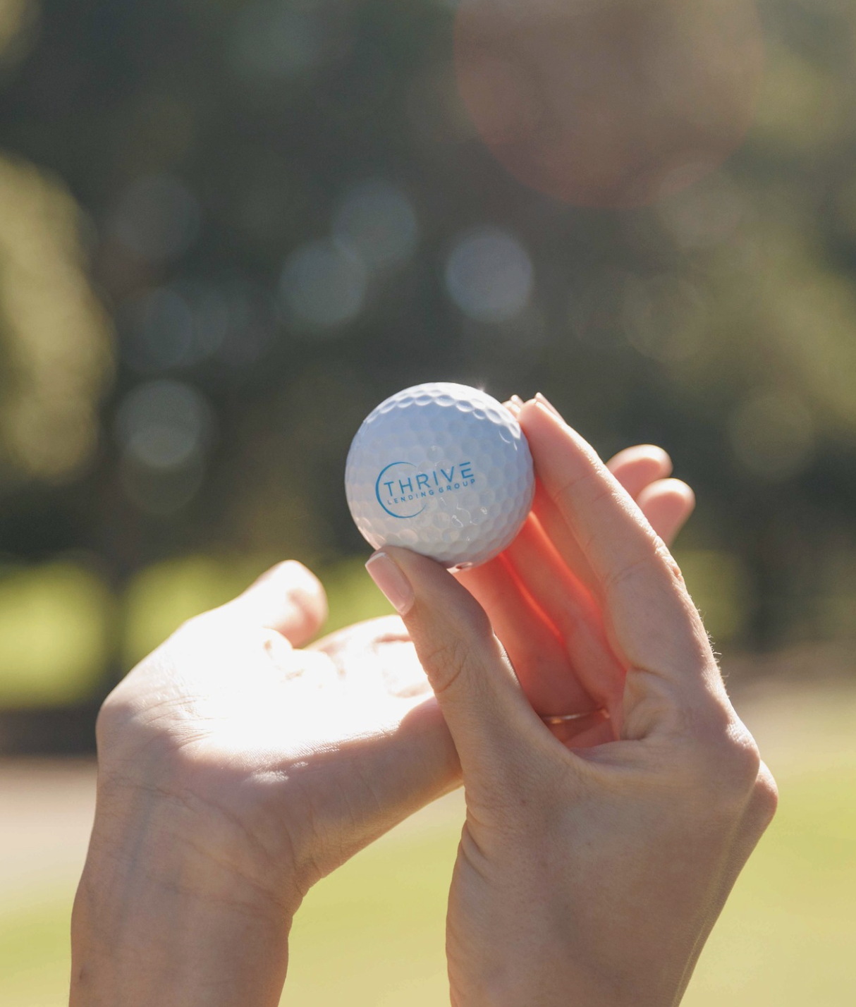A person holding a golf ball with the logo 'Thrive Lending Group' on it, outdoors with a blurred background of trees and sunlight.