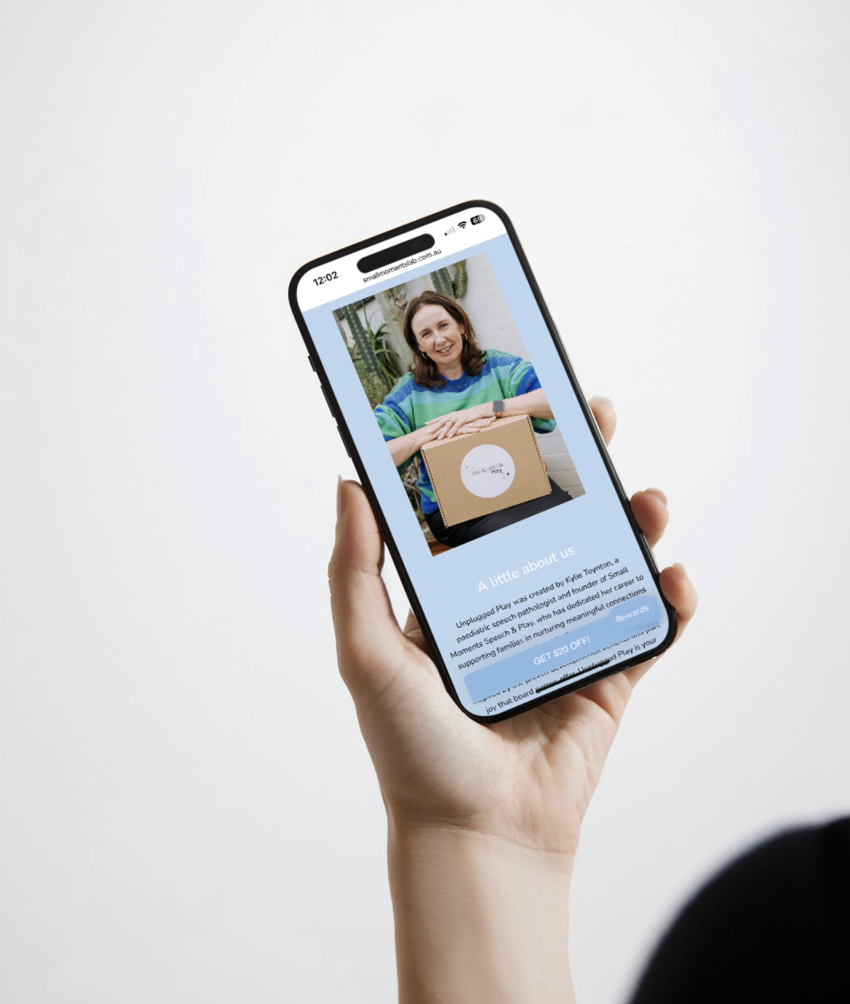 A person holds a smartphone displaying a webpage with a woman sitting at a table, smiling, wearing a colorful striped shirt, with a cardboard box labeled 'Unplugged Play' in front of her, against a background of indoor plants.