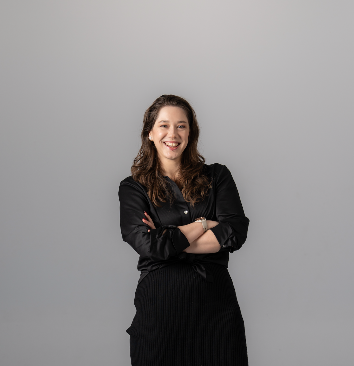 A smiling woman with brown hair, wearing a black satin shirt and black skirt, standing against a plain grey background.
