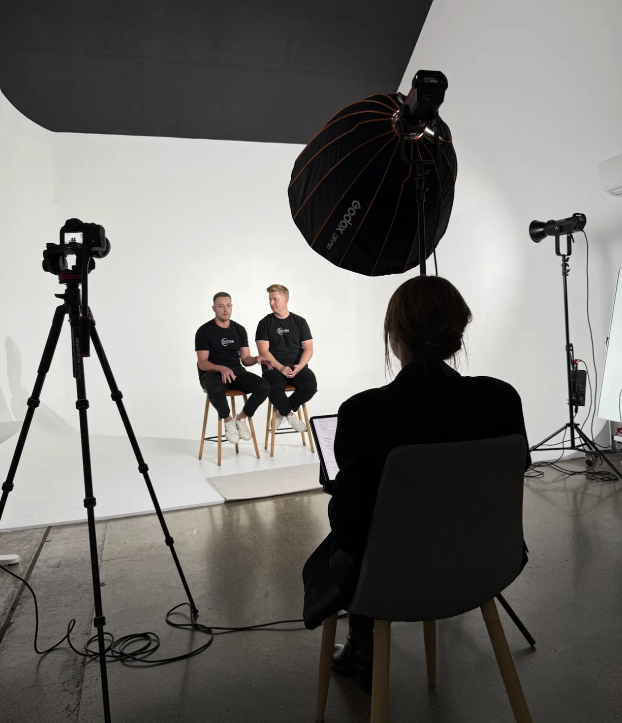 Two men sit on stools in a photography studio with a white background, being filmed by a woman using a laptop, surrounded by professional lighting and camera equipment.
