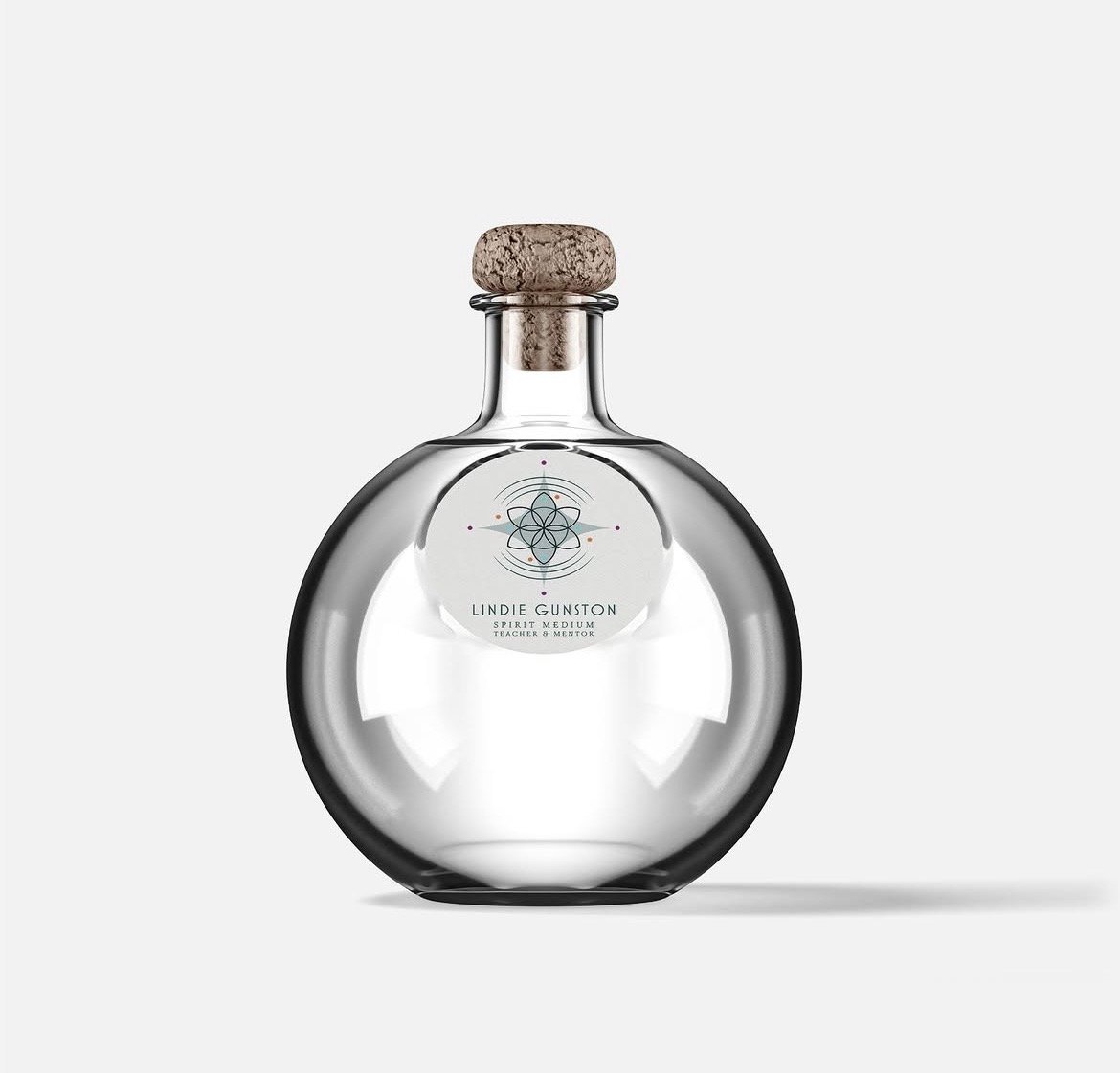 A round glass bottle with a cork stopper, featuring a geometric design and the name "Lindie Ginston" on the label, indicating it is a spirit medium.