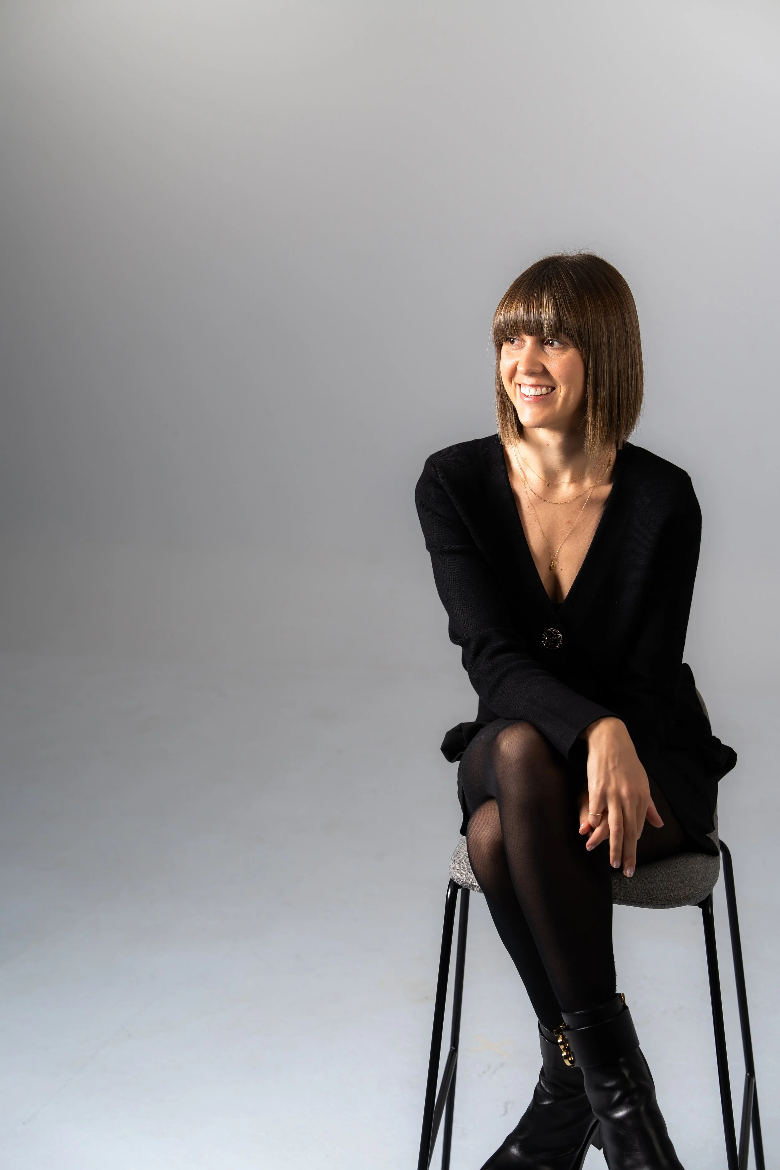 A woman with shoulder-length brown hair and bangs sitting on a chair against a light gray background, smiling and looking to her right.