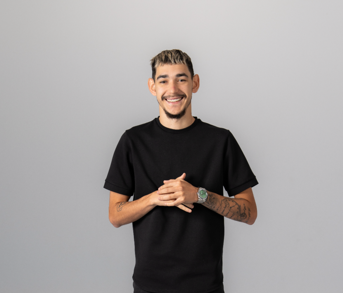 Smiling young man with short hair and tattoos on forearms, wearing a black t-shirt, standing against a plain light gray background.