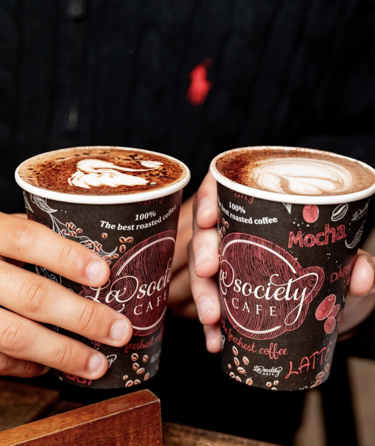 Two hands holding cups of coffee with latte art on top, featuring a heart and leaf design, against a dark background.