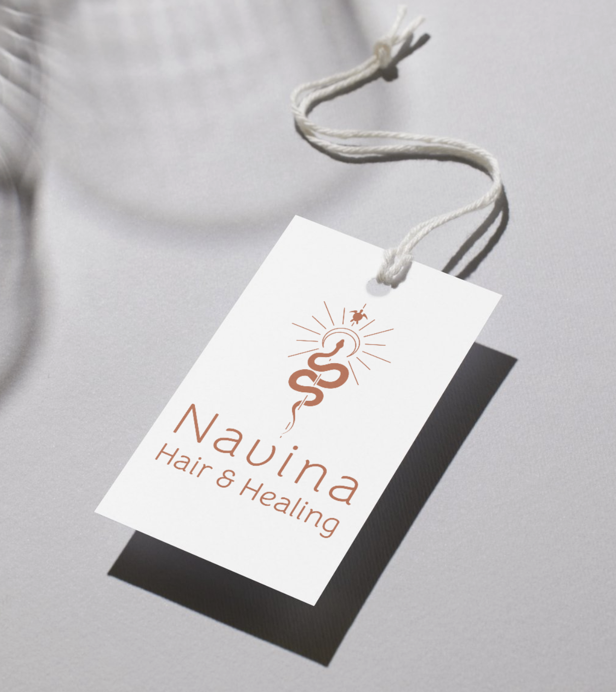 A white tag with the logo of a snake, sun, and insect, attached to a string, with the text "Nauina Hair & Healing" in brown, lying on a gray surface.
