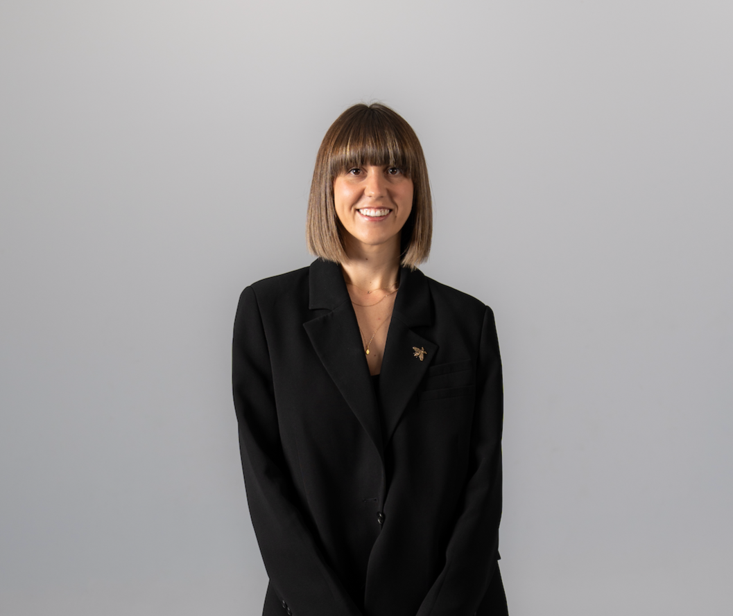 A woman with shoulder-length brown hair and bangs, smiling, dressed in a black blazer with a pin, standing in front of a plain light gray background.