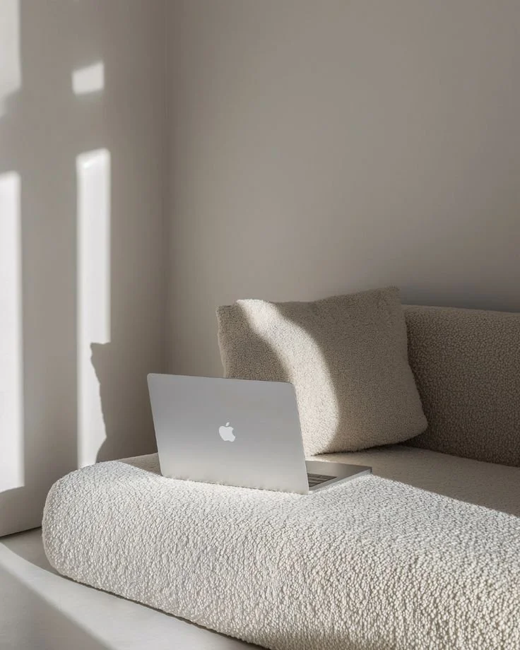 A silver Apple MacBook on a textured beige cushion on a beige sofa with a matching pillow, with sunlight casting shadows on a white wall.