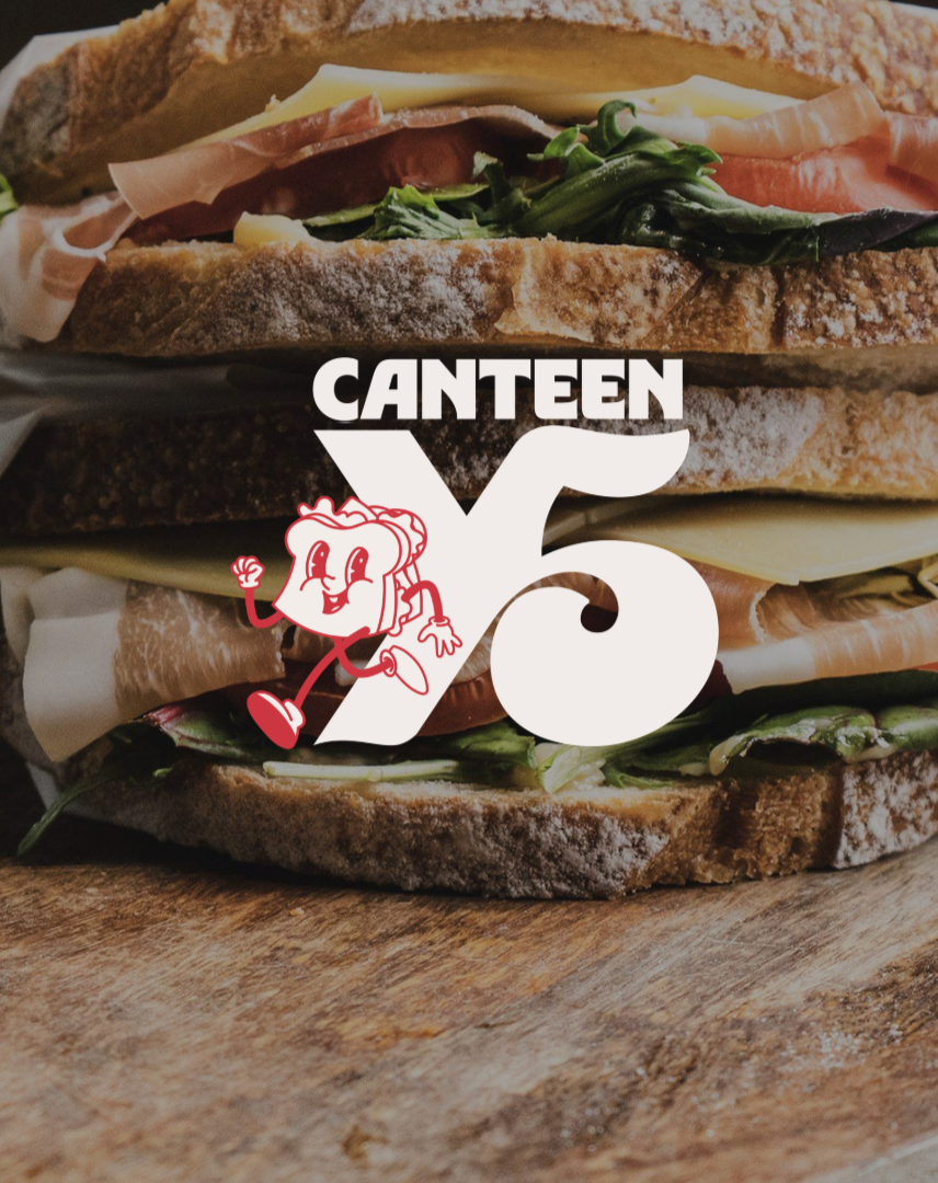 Close-up of a sandwich with whole grain bread, cheese, ham, smoked salmon, and greens on a wooden surface, with a Canteen Valley 76 logo overlay.