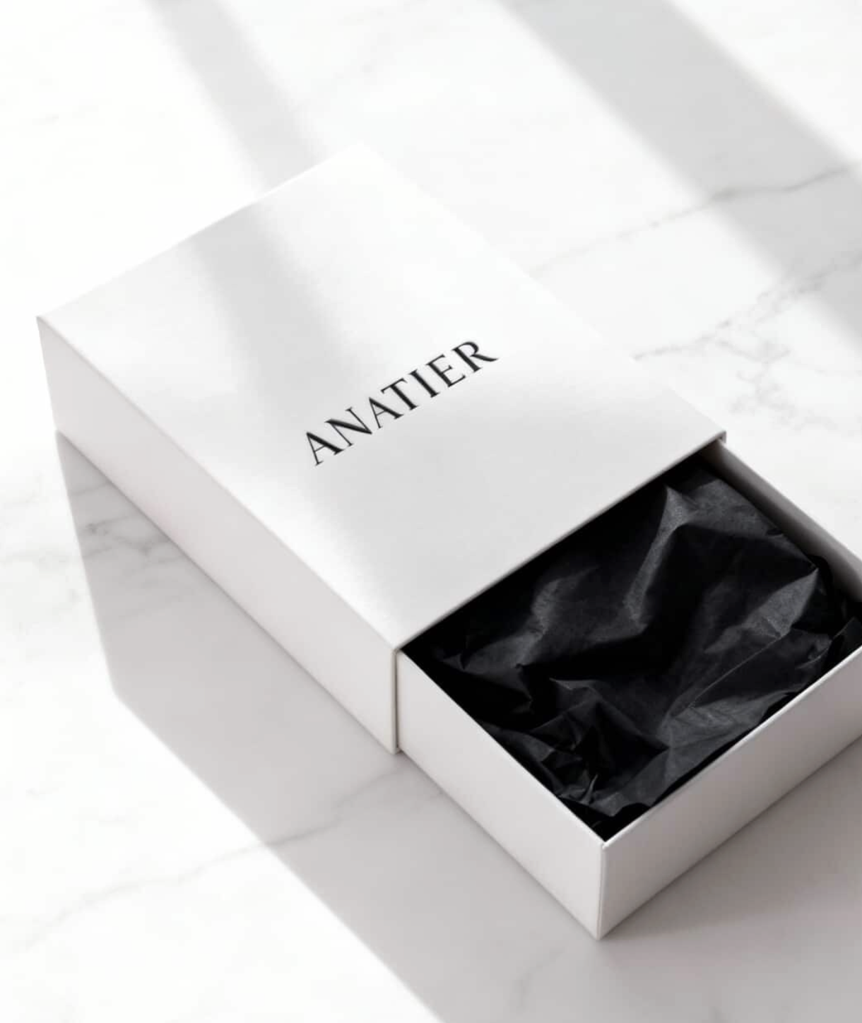 Open white box with the word 'ANATIER' printed on the lid, black paper lining inside, on a marble surface.
