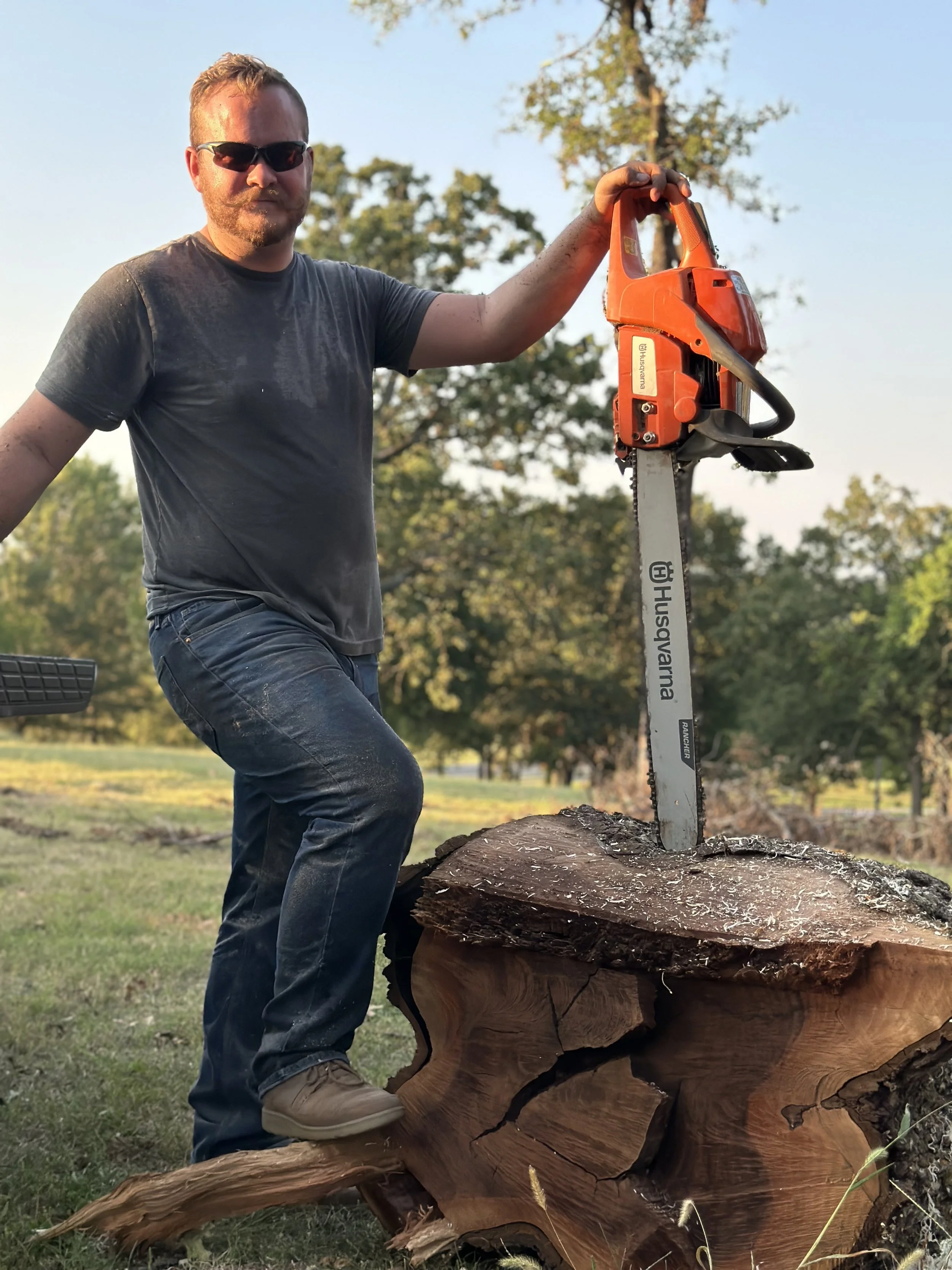 A man wearing sunglasses and a dark t-shirt stands outdoors, holding a chainsaw upright with one foot on a large tree stump.
