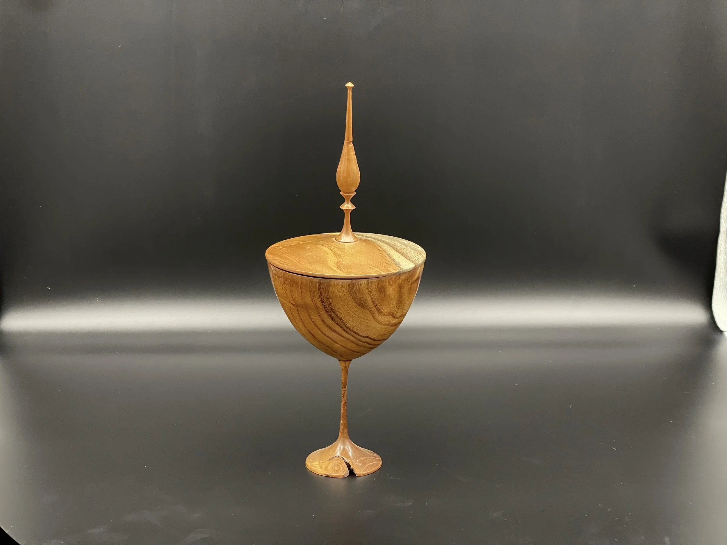 Wooden decorative object resembling a goblet with a tall, slender spire on top, set against a black background.