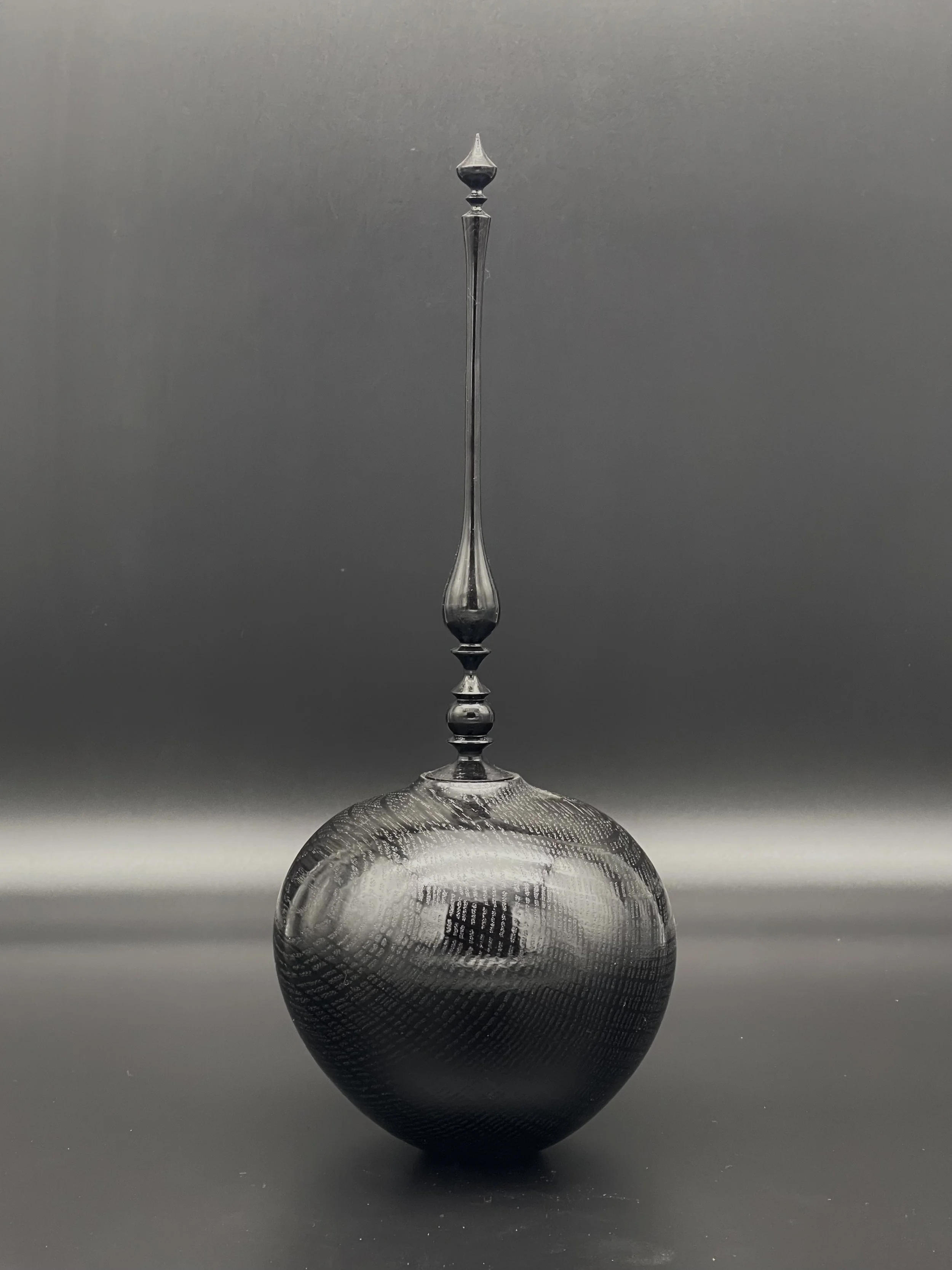 A tall, hollow vessel with a finial with decorative elements extending from a round, textured black wood against a black background.