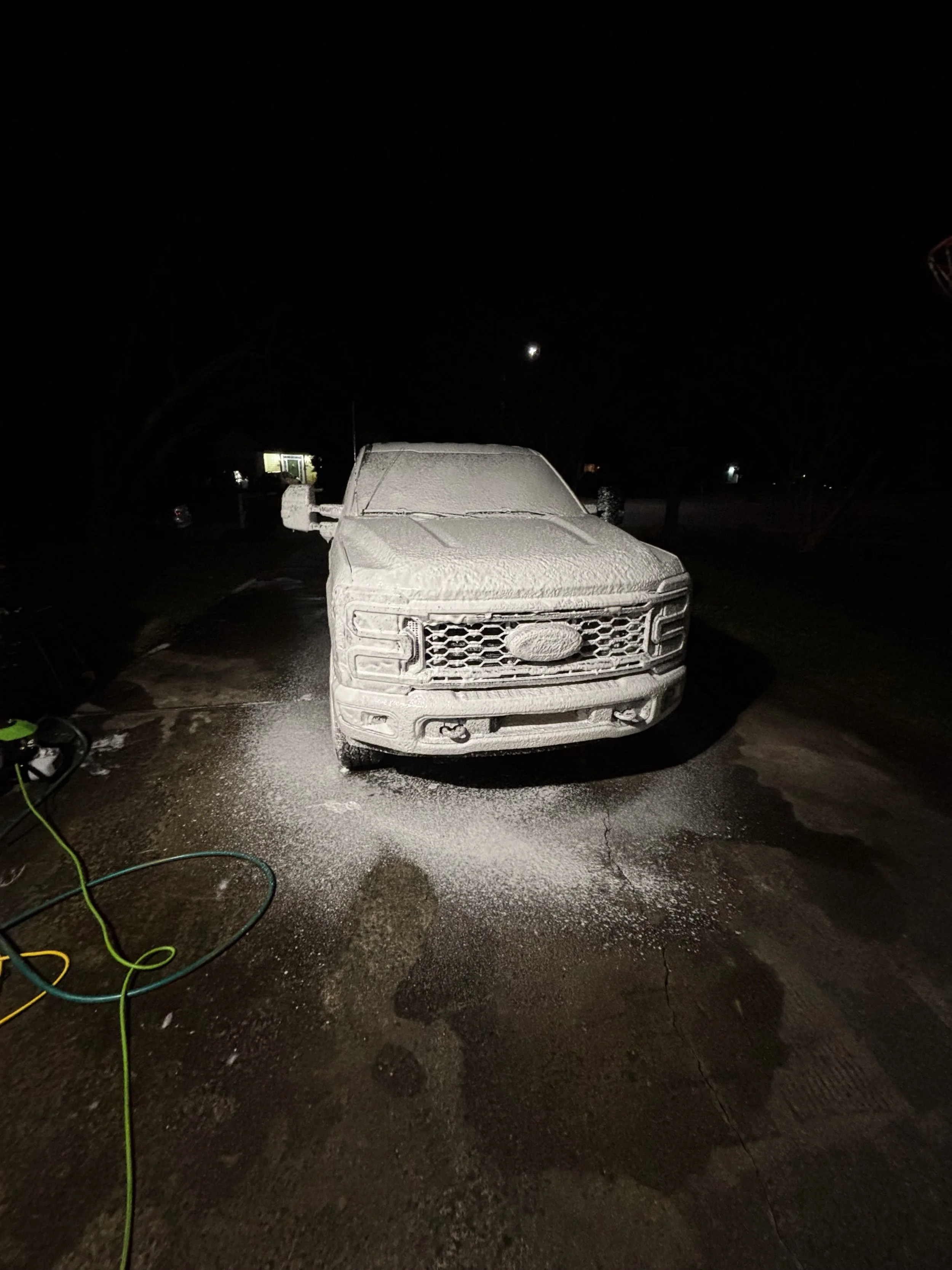 A white pickup truck covered in soap suds during a nighttime car wash, with a hose on the ground nearby.