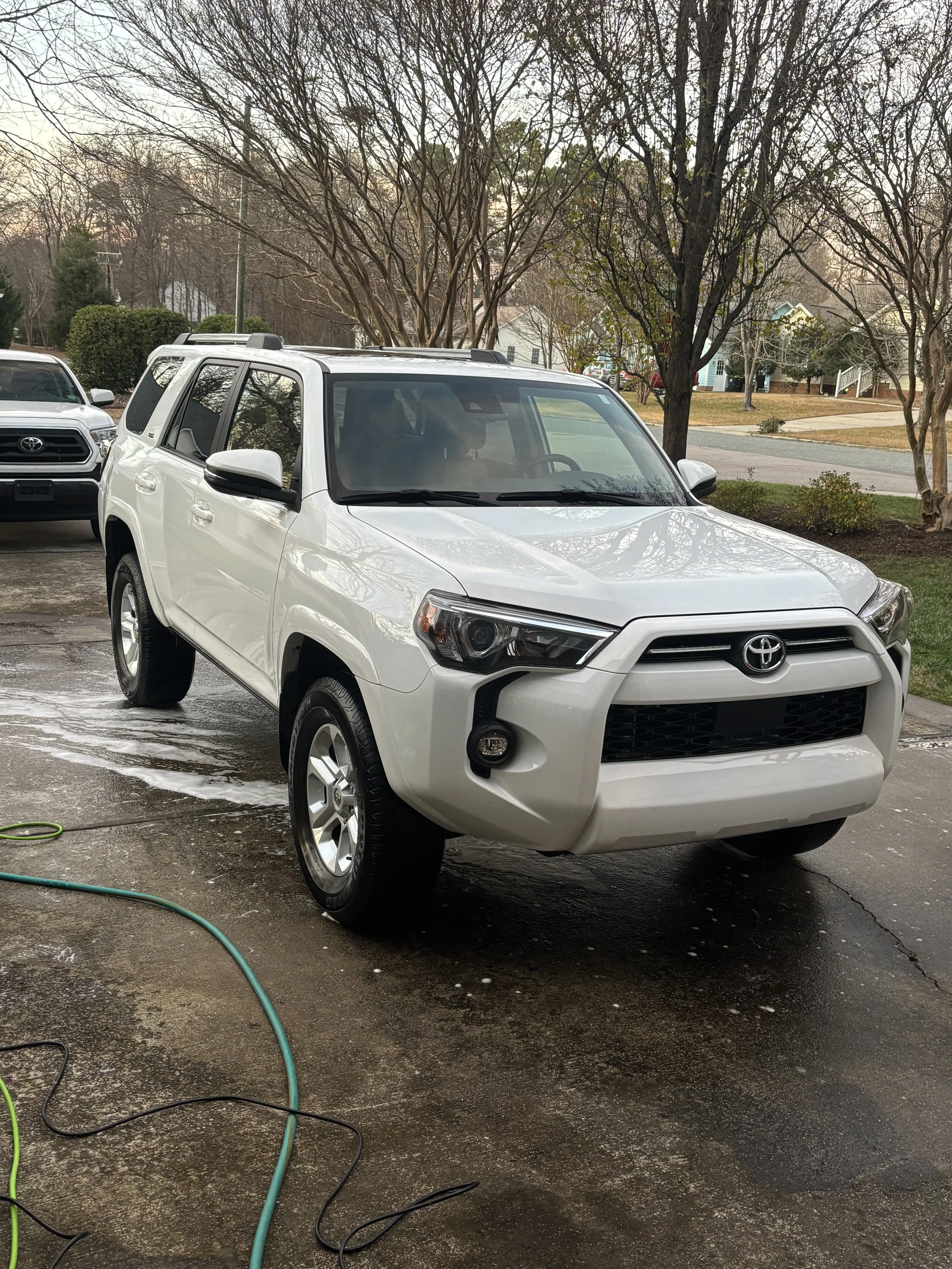 White Toyota SUV being washed outdoors with soap and water on a driveway, with another vehicle and residential neighborhood in the background.