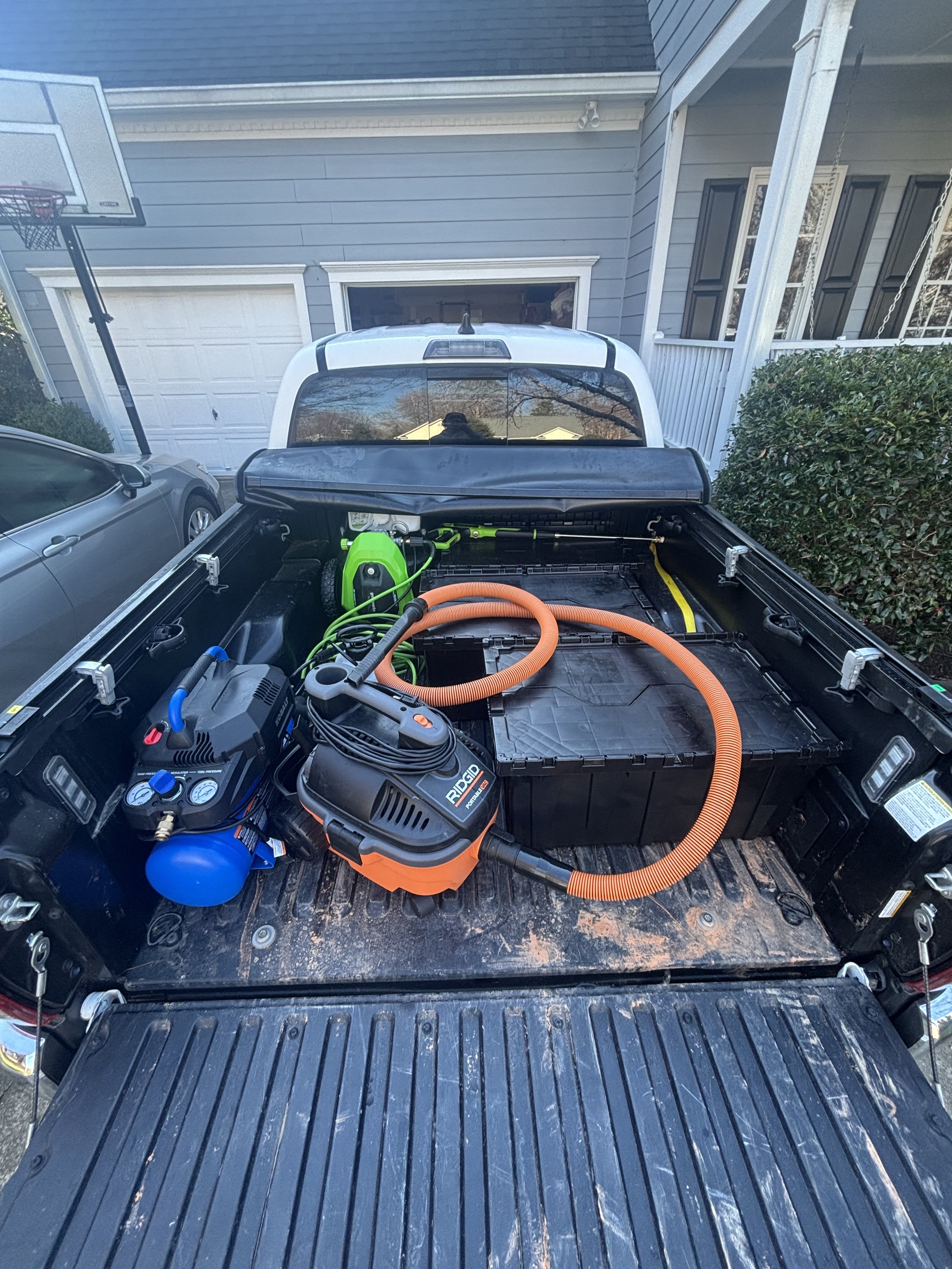 The back of a truck showing equipment including a portable air compressor, a leaf blower, and a large black storage container, with a house and a basketball hoop in the background.